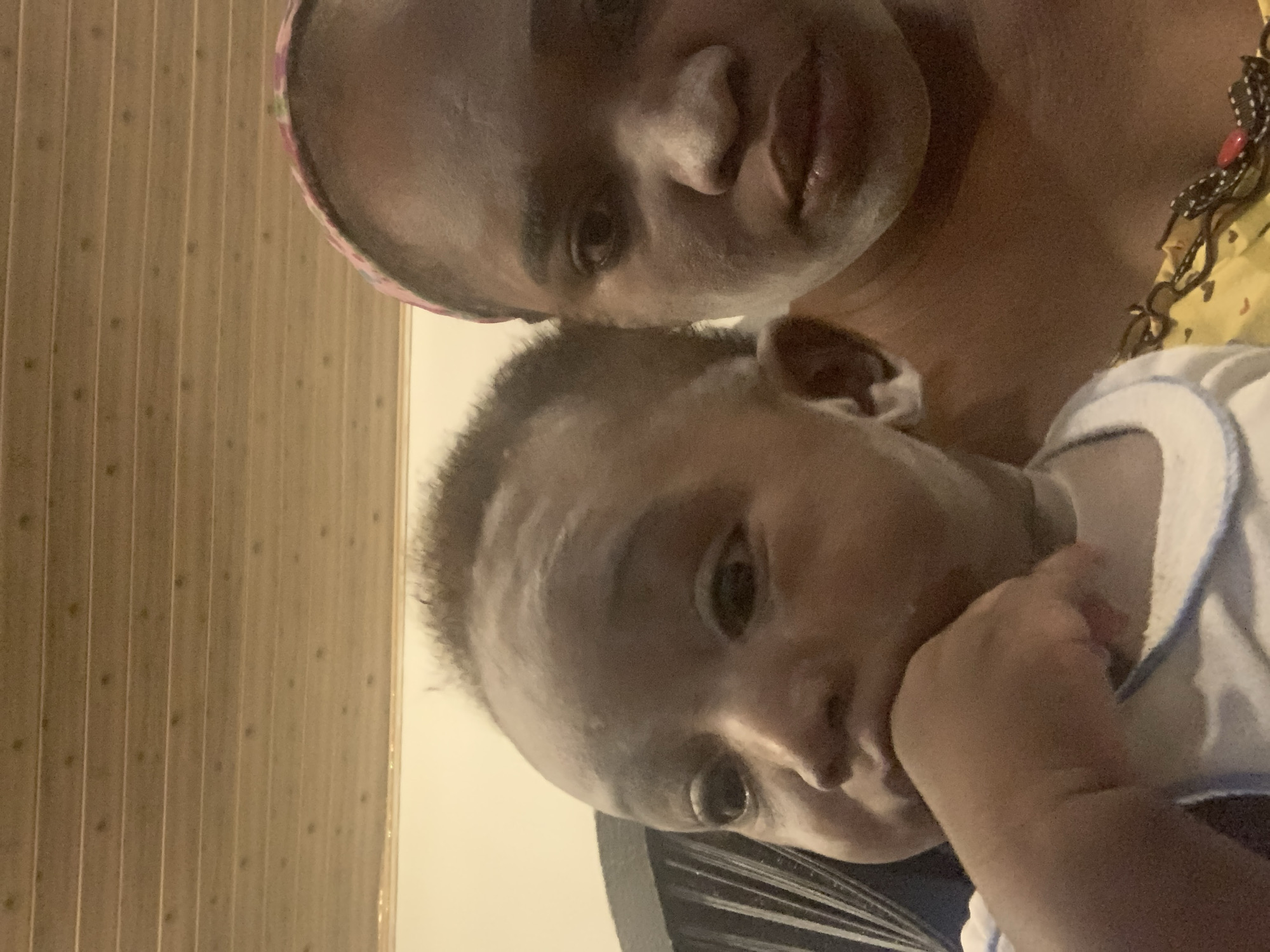 An overhead, close-up shot depicts an adult and a baby. The adult appears to be holding the baby. The baby is partially visible, with their face, one hand, and part of the torso showing. The baby is wearing a light-colored shirt with a contrasting trim. The adult is positioned behind the baby, with their head and part of their shoulder visible. The adult is wearing a headband. The background includes a wall with vertical wooden panels. The location is assumed to be Barakin Akawo, Nigeria.