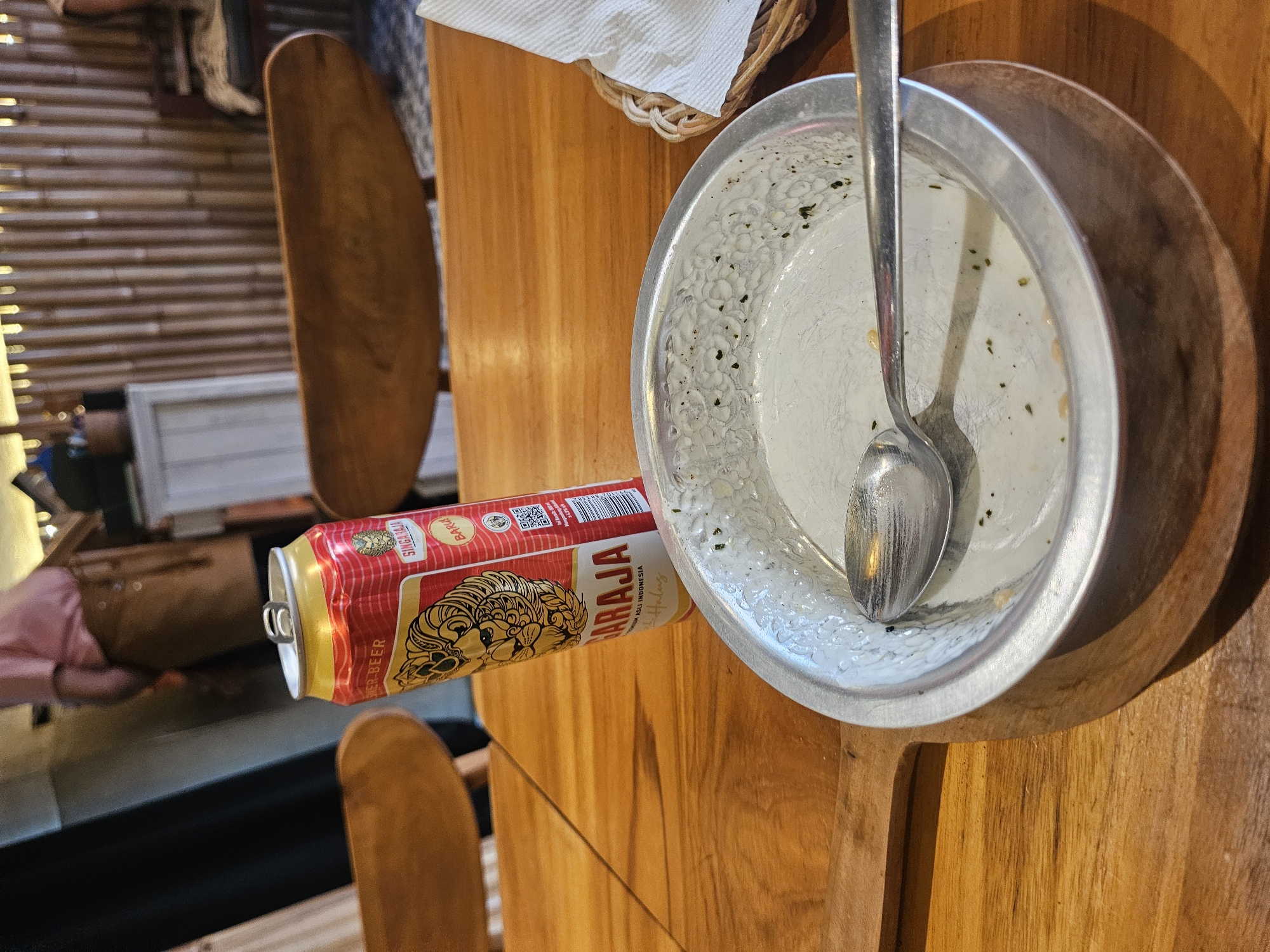 A Singaraja beer can and a used metal bowl with a spoon are on a wooden table. The bowl contains remnants of food. A person in a pink shirt and brown apron is visible in the background, as are bamboo walls and wooden chairs. The scene appears to be an indoor dining area, likely a restaurant or cafe in the Ubud District of Indonesia.