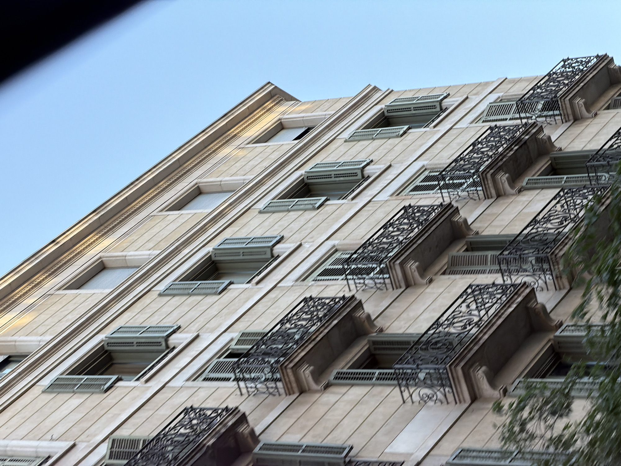 This image shows the exterior of a multi-story building with a light-colored facade, likely made of stone or stucco, in Barcelona, Spain. The building features rows of windows with pale green shutters, some of which are open. Several balconies with ornate wrought-iron railings extend from the facade. The architecture suggests a style common in the early to mid-20th century. The sky above is a clear blue, indicating daytime. The bottom portion of the image is partially obscured by blurred green foliage, possibly trees or bushes growing in front of the building. No people are visible in the frame.