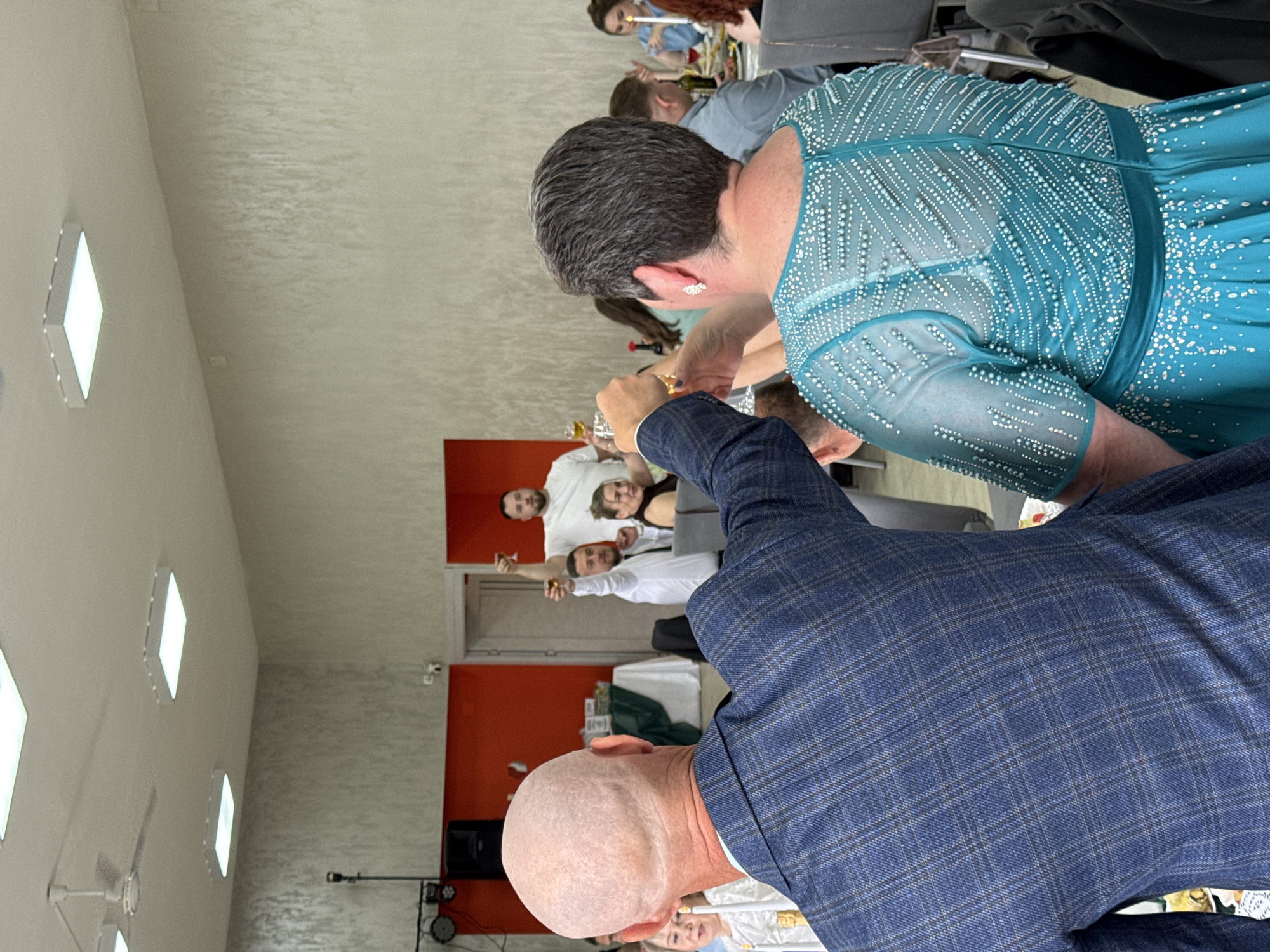 This image captures an indoor celebratory gathering, likely a party or reception, in Chaikovsky, Russia. The perspective is from behind two individuals in the foreground, looking towards a group of people in the background.
In the immediate foreground, the back of a person with a bald head is visible, wearing a blue plaid suit jacket. To their left, the back of a person with short, dark grey hair is seen, adorned in an elaborate teal dress with sheer sleeves and intricate white beading across the bodice and sleeves. Both appear to be holding up glasses, participating in a toast or similar celebratory action.
In the middle ground and background, several other individuals are visible, some seated at tables. Many are looking towards the camera and raising their glasses, smiling, indicating a shared moment of celebration. One person in the background is wearing a white t-shirt and holding a glass.
The setting appears to be a well-lit event space or banquet hall. The walls are light-colored, with a distinctive bright orange accent wall in the background. The ceiling features multiple modern, diamond-shaped light fixtures and at least one ceiling fan. Other visible objects include tables, and what appears to be sound equipment or speakers against the orange wall. The overall mood conveyed by the participants' expressions and actions is joyful and festive. No visible text is present in the image.