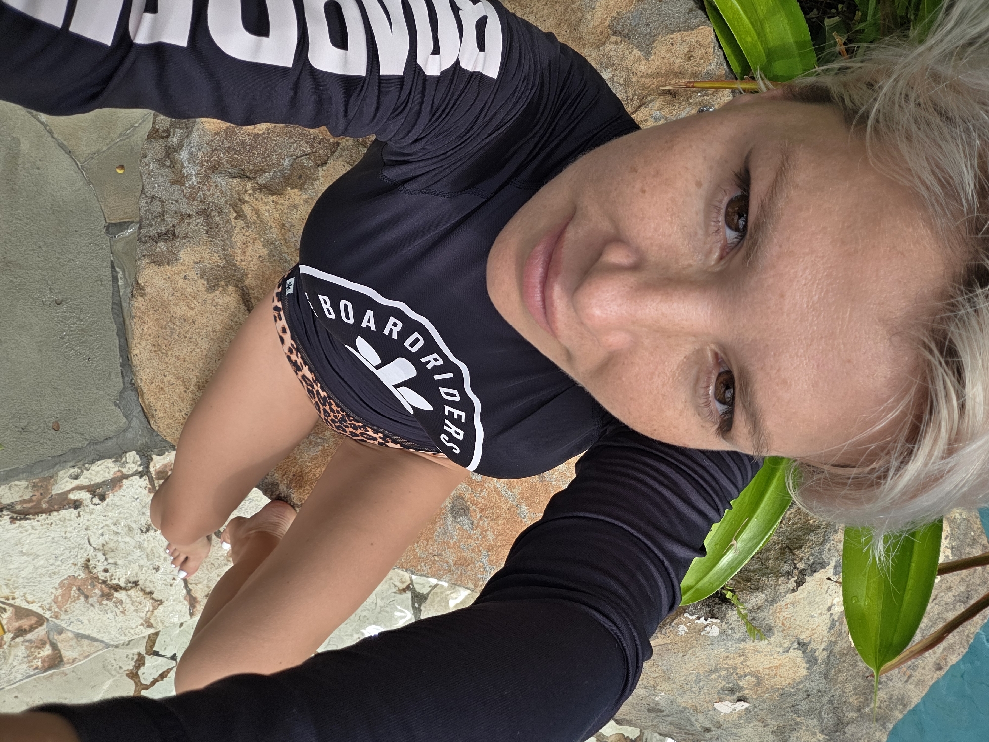 A selfie depicts a person with fair skin, short blonde hair, and freckles, wearing a black long-sleeved shirt with "BOARD RIDERS" printed on the front. The person is positioned at an angle, looking directly into the camera. The background consists of a stone surface, possibly part of a pool area, and greenery. The location is in Kuta, Indonesia. The person's legs and feet are also visible. The person's toes have white nail polish.