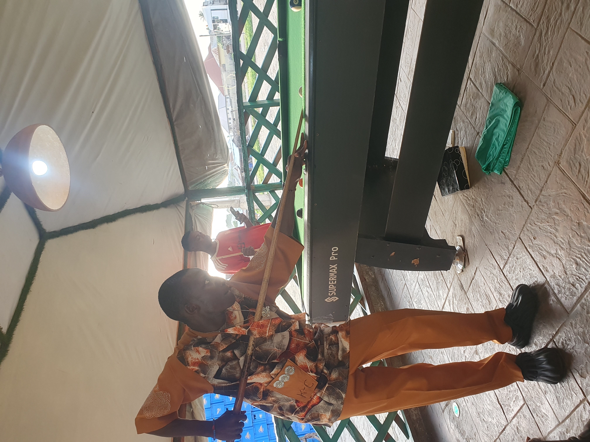 In Lokoja, Nigeria, a person is playing pool. The scene takes place outdoors under a white tent-like structure. The main subject, a Black male wearing an orange and brown patterned shirt and orange pants, is in the foreground, focused on his shot. He is holding a pool cue and is positioned behind the pool table. Another Black male, wearing a red shirt, is visible in the background, looking at his phone. The pool table is dark green with a "SUPERMAX Pro" logo. The floor is paved with textured tiles. A green cloth is folded on the ground near the pool table. The environment suggests a relaxed, recreational setting.