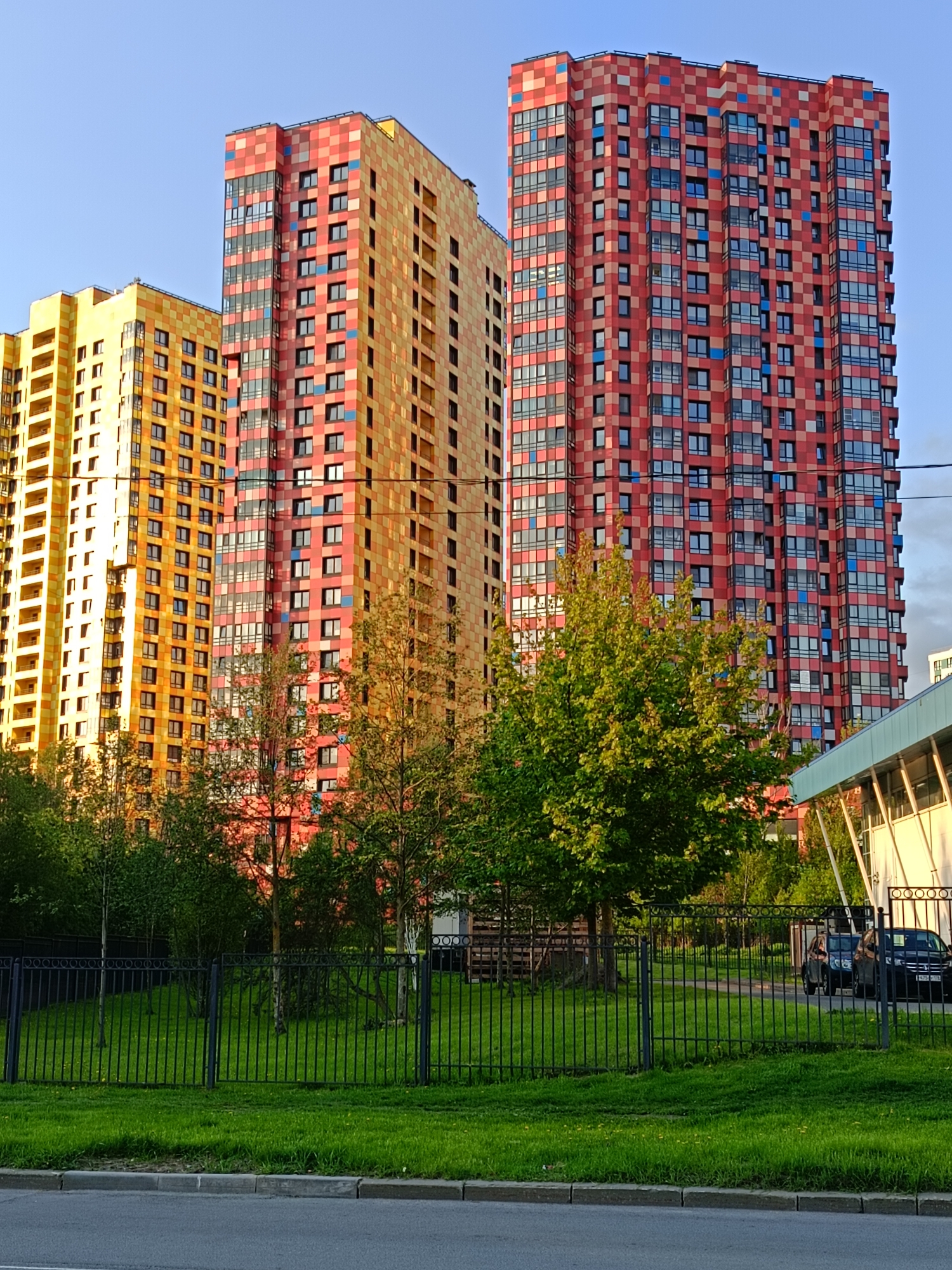 The image shows three high-rise apartment buildings in Saint Petersburg, Russia. The buildings are characterized by their checkerboard facades in yellow and red hues. Green trees and manicured lawns are visible in front of the buildings, separated by a black metal fence. Two cars are parked in front of one of the buildings on the right. The sky is blue and bright. A building with a white wall and slanted roof is on the right side of the image. The photograph appears to be taken from a street-level vantage point.