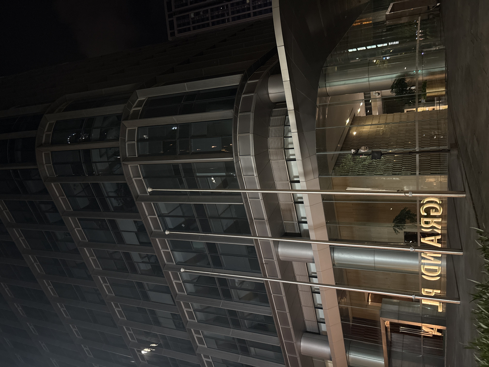 The image depicts the exterior of a modern, illuminated building at night. The building features a sleek, contemporary design with extensive glass facades and metallic accents. Horizontal bars made of a reflective material run across the front of the building, acting as architectural elements.
Reflections on the glass reveal an indoor setting with stairs and greenery. Visible text on the building's facade reads "GRAND BL." in large, illuminated golden letters, with the letters appearing reversed due to reflection. The scene is set in an urban environment, likely a commercial or business district, given the architecture and signage.
The time of day is clearly night, indicated by the darkness and artificial lighting. There are no people visible in the image, and the focus is on the architectural structure and its illuminated signage. The overall impression is one of a well-lit, modern establishment. The location context of Haizhu District, China, suggests this is likely a significant building within that area.