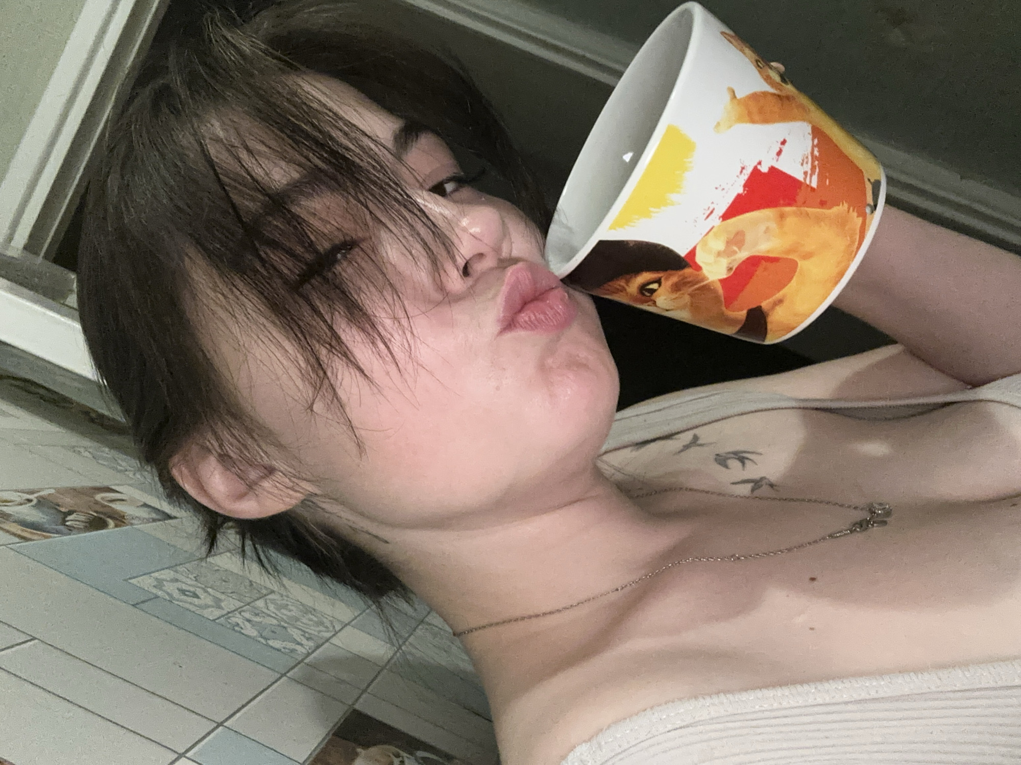 A young woman with short, dark hair is holding a white mug with a colorful cat design. She is puckering her lips and looking towards the camera. She is wearing a light-colored, ribbed tank top and a silver necklace. There are tattoos visible on her upper chest and left shoulder. The background appears to be a bathroom with tiled walls and a partially visible doorframe. The lighting suggests an indoor setting, possibly at night or with dim artificial light. There is no visible indication of the specific location being in Chaikovsky, Russia.