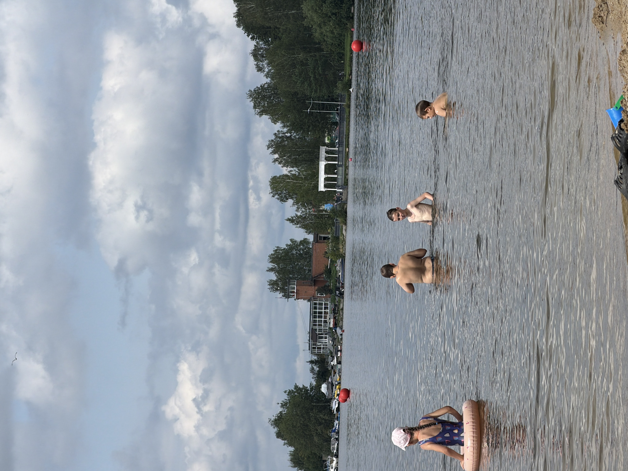 The image depicts a recreational scene on a body of water, likely a lake or river, on a cloudy day. In the foreground, a young girl with a pink hat and a swimsuit is sitting on a pink inflatable ring. Further back in the water, four individuals are visible. Two are adult males, one facing away from the camera and another in the distance. Two young boys are also in the water. In the middle ground, a building with a tower and a colonnaded structure are visible on the shore, surrounded by trees. Several boats are docked along the shore. Two large red buoys are in the water. The foreground also shows a sandy bank with some toys. The overall impression is a summer day at a public swimming area. The location is identified as Zarechny, Russia.