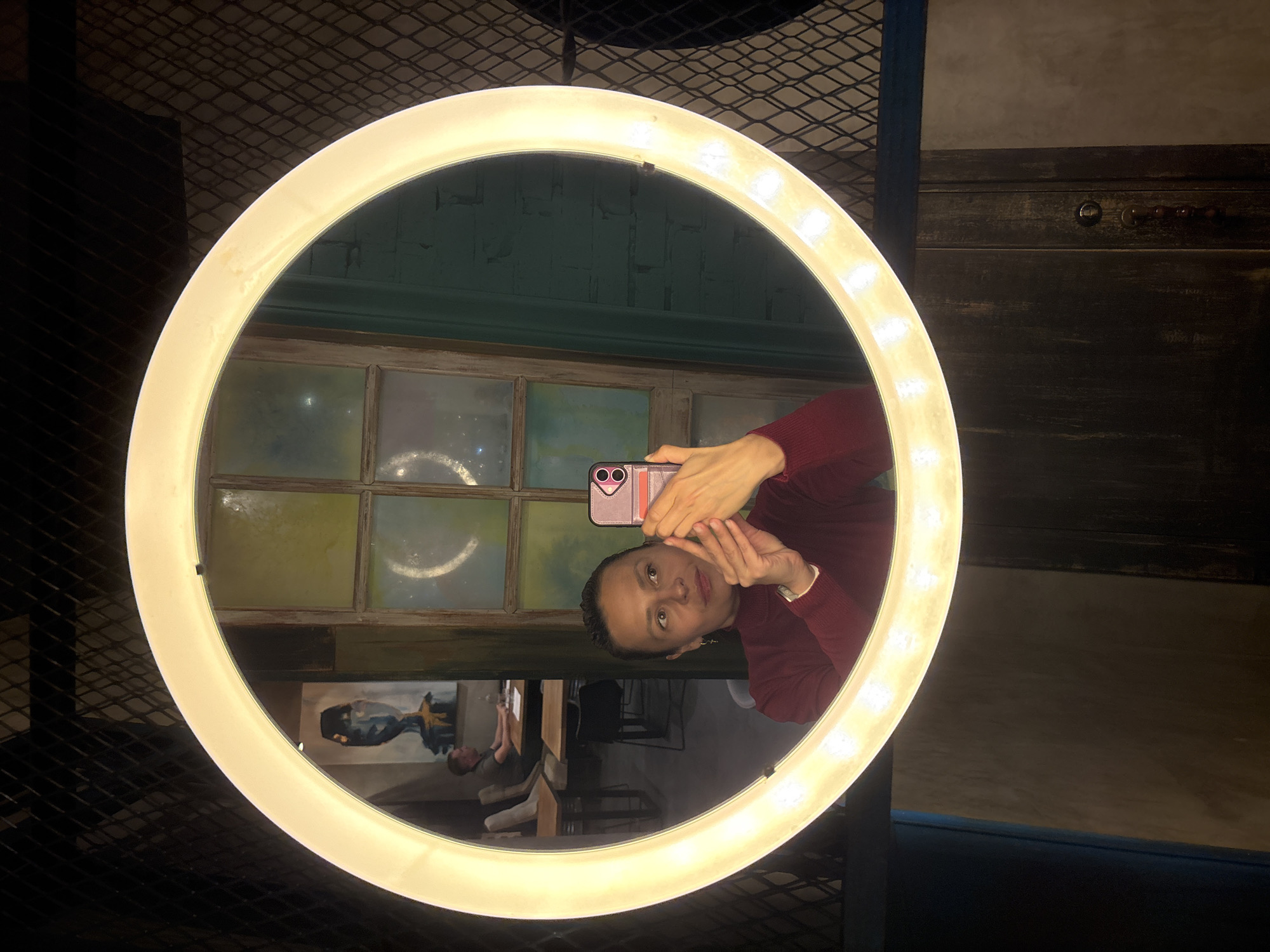 A woman takes a selfie using a ring light, with her reflection visible in a mirror. The scene appears to be inside a cafe or restaurant in Kazan, Russia, based on the Cyrillic writing on the windows and overall interior design. There is dark metal lattice work on the left side of the frame. A blurred painting or mural can be seen in the reflection, along with tables, chairs, and another person in the background.