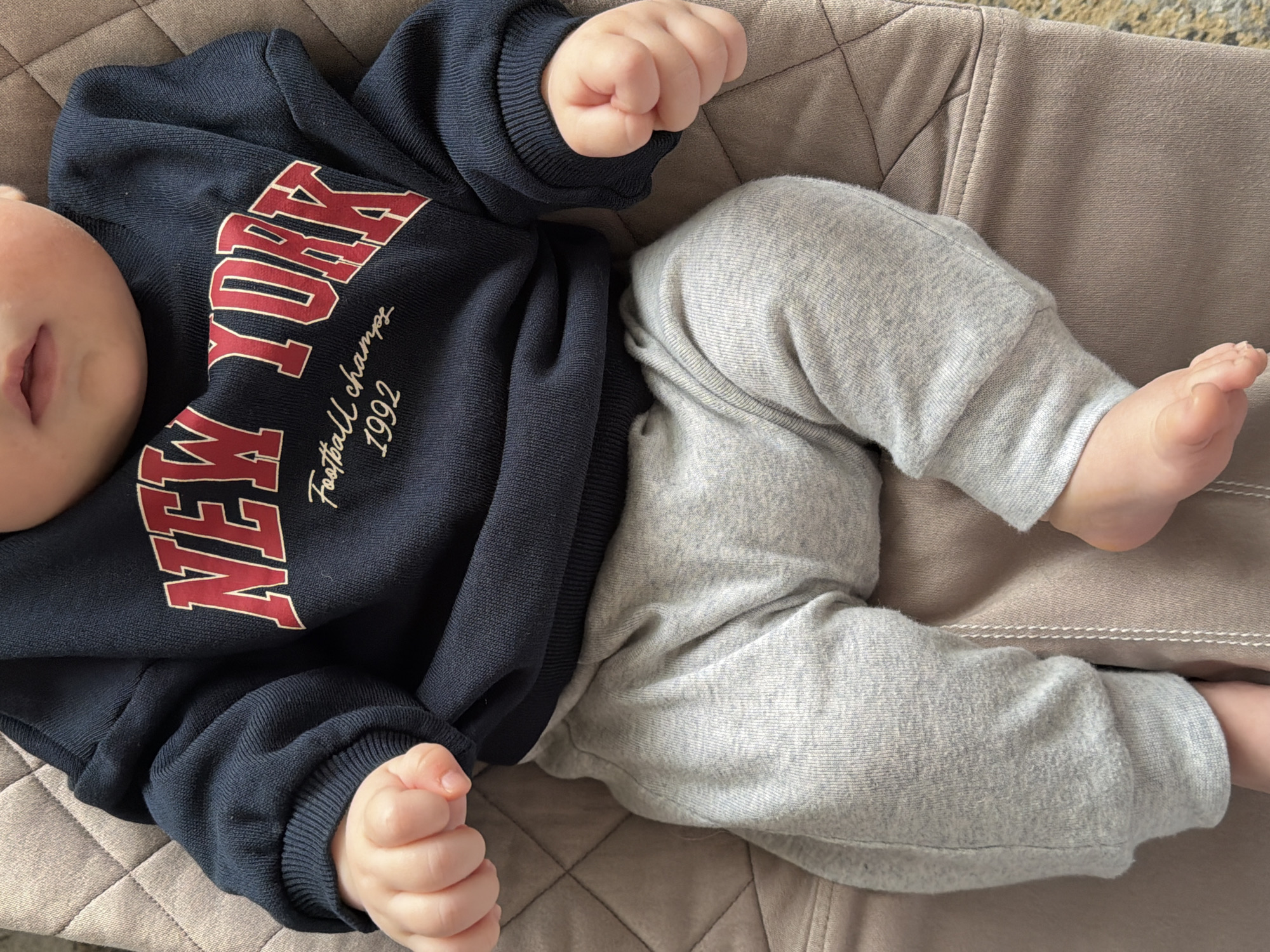 An overhead shot captures a baby lying comfortably on a beige quilted surface, likely a bouncer or a soft chair, in an indoor setting. The baby is dressed in a navy blue long-sleeved sweatshirt and light grey sweatpants. The sweatshirt features bold red text with a white outline that reads "NEW YORK" across the chest. Below this, in a smaller, cursive font, the words "Football champs" and "1992" are visible. The baby's head is partially seen on the left, with a glimpse of their mouth. Both of the baby's hands are curled into gentle fists, resting near their body. Their legs are bent, indicating a relaxed posture. The image suggests a peaceful moment, with the baby appearing calm.