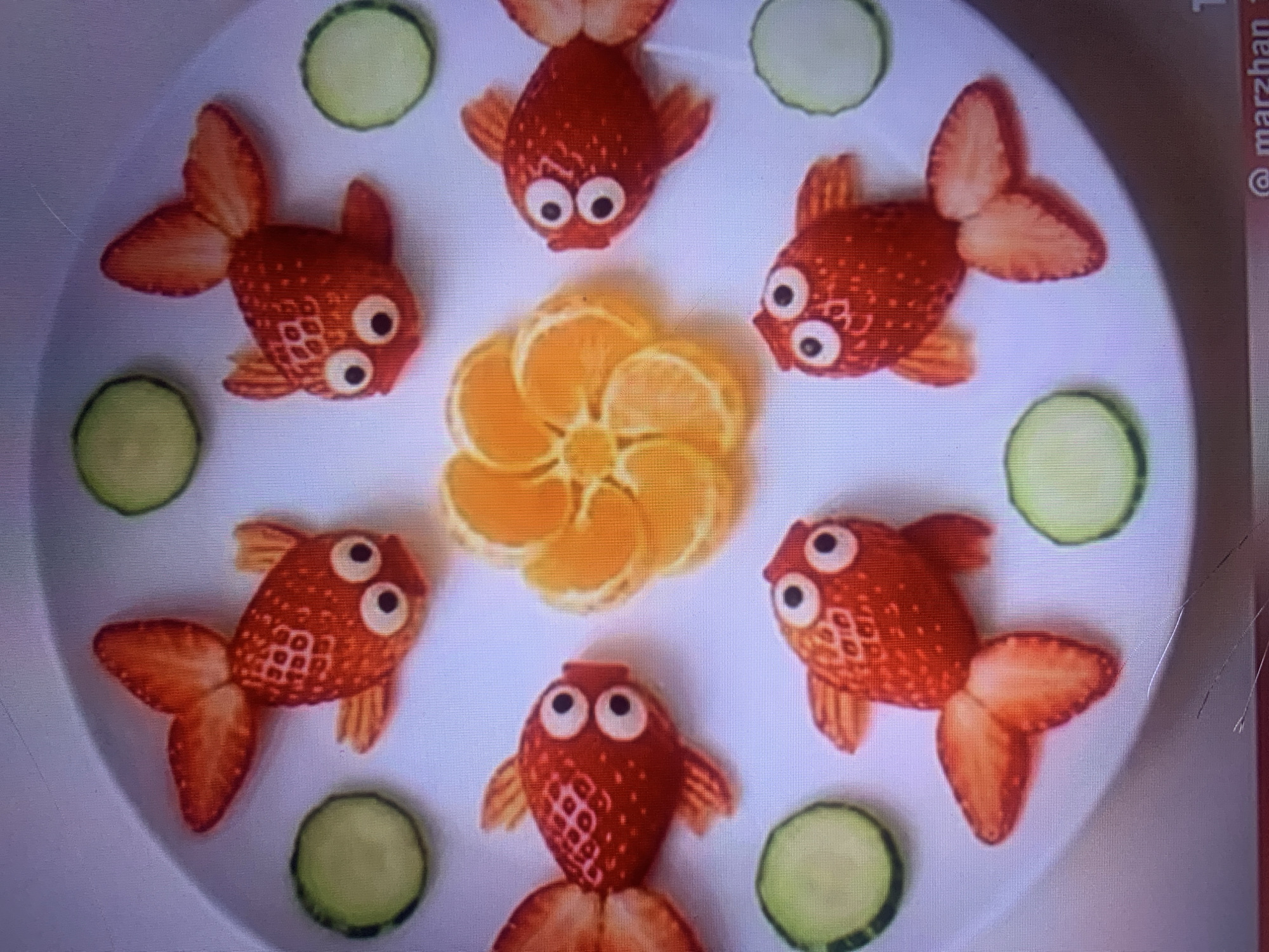 The image shows a white plate filled with fruit arranged to resemble goldfish and a flower. There are six strawberry goldfish, each with cucumber slices for eyes and orange slices for fins and tails. In the center of the plate, orange slices are arranged in a flower shape. Around the goldfish and flower, there are several slices of cucumber. The background is blurred, but appears to be a textured surface. The scene is likely a still life arrangement, possibly for a food presentation or a creative project. There is no indication of the city of Almaty, Kazakhstan, or any specific location within it.
