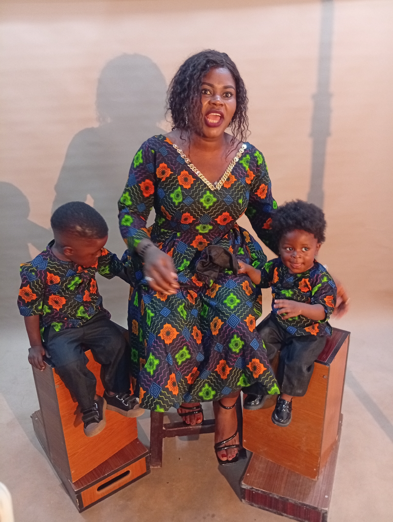 A Black woman with dark, curly hair and an open mouth is seated on a wooden stool in front of a tan wall, wearing a long-sleeved, colorful dress with an orange, green, and blue geometric pattern and a gold trim neckline. To her left, a young Black boy in a matching patterned shirt and dark pants sits on a wooden box, looking down. To her right, a toddler Black boy in a similar patterned shirt and dark pants sits on another wooden box, looking to the side and sticking his tongue out. The woman has her left hand resting on the toddler's back. The lighting casts a shadow of the woman on the wall behind her. The scene appears to be in a studio with wooden platforms and boxes.