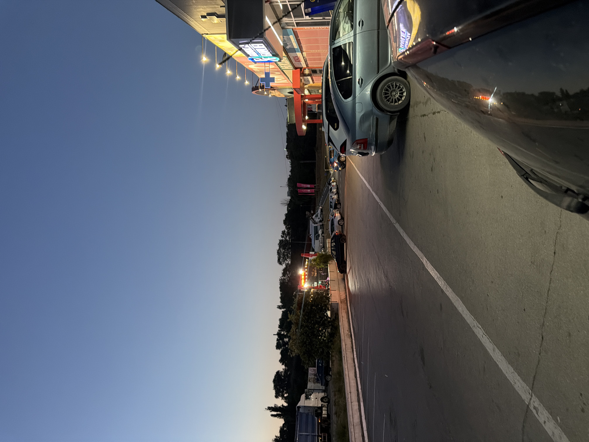 This is a vertical, eye-level, full shot taken at dusk in Orhei, Moldova. In the foreground, the hood of a dark-colored car is visible, reflecting some red and yellow lights. To the left and slightly behind it, a light blue sedan is parked. In the midground, a paved road stretches into the distance, bordered by a white line on the left and a row of parked cars. Further back, a line of trees forms a dark silhouette against the twilight sky. A brightly lit building with signs and exterior lights runs along the right side of the frame. In the distance, several trucks and white vans are parked in a lot. The sky above is a clear, deep blue, transitioning to a lighter hue near the horizon.