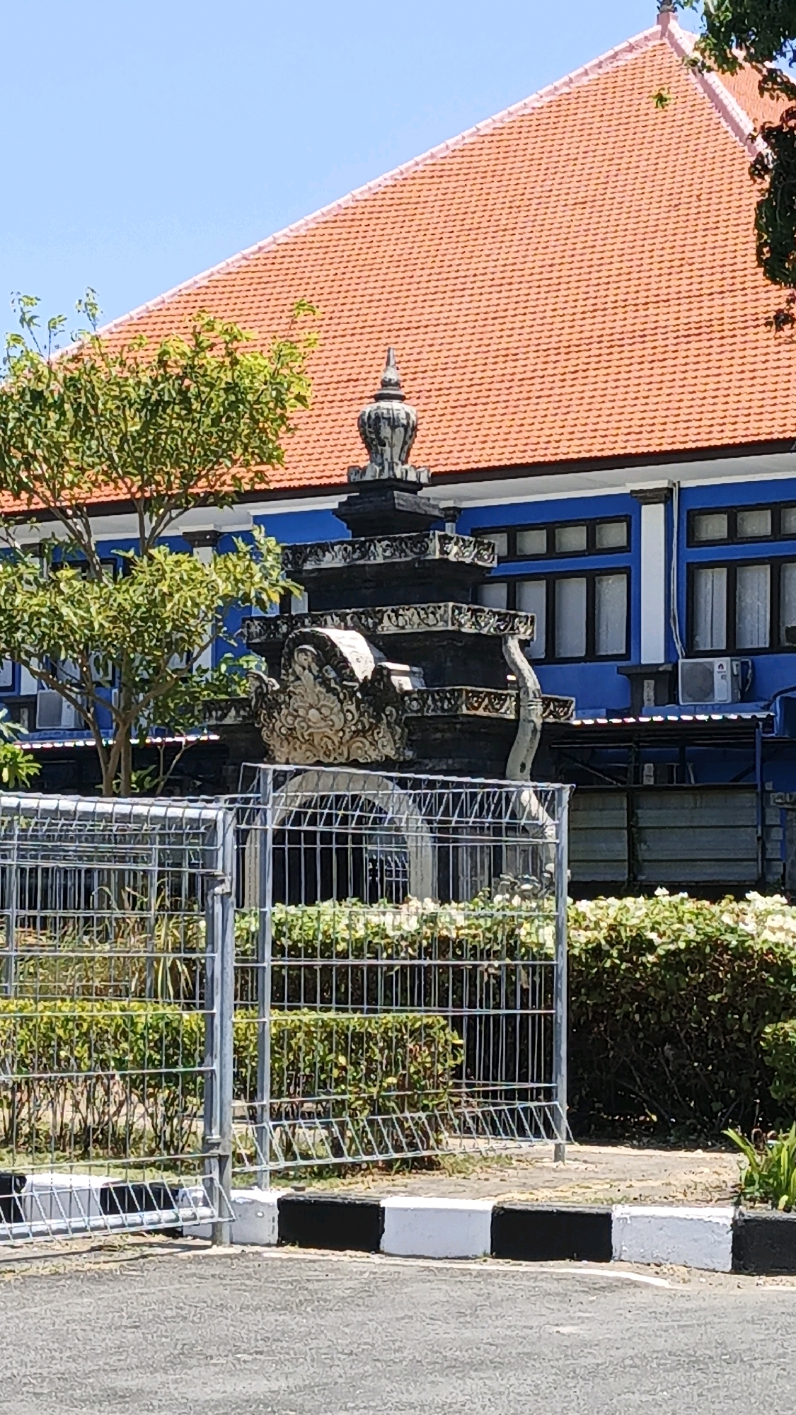 The image depicts an outdoor scene on a clear, sunny day, focusing on a building with a red tiled roof and blue exterior walls. In front of the building, a decorative stone structure, possibly part of a gate or monument, is visible, featuring intricate carvings. A metal fence runs across the foreground, with lush green bushes and some flowering plants behind it. To the left of the stone structure, a leafy green tree stands tall. The building has several windows and what appear to be air conditioning units mounted on the exterior. The scene suggests a location within a city, likely a public or institutional space, given the presence of the fence and the architecture. Without specific identifying markers, pinpointing the exact location in Denpasar, Indonesia, is not possible, but the architectural style and vegetation are consistent with the region. There are no people visible in the image.