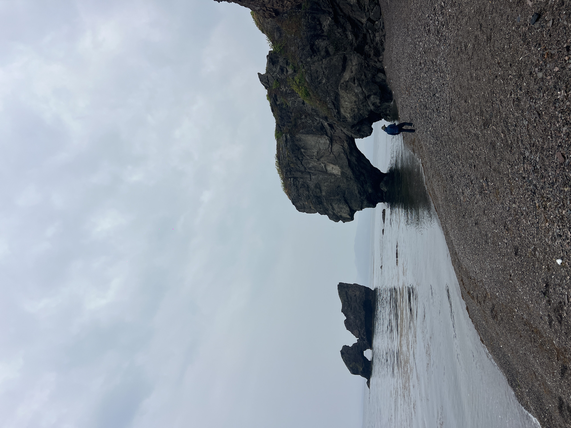 The photograph shows a beach in Gvozdevo, Russia. The sky is overcast. In the center of the frame are two rock formations with natural arches. A person wearing a blue jacket is standing near the base of the larger rock formation. The beach is composed of dark-colored pebbles. The water is reflecting the sky.