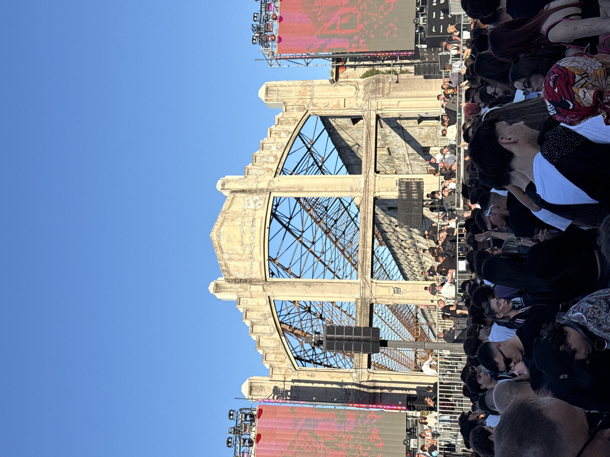 The image captures a large crowd of people gathered in what appears to be an outdoor event or concert setting, with a prominent architectural structure in the background. The architectural structure has a skeletal frame, possibly an old factory building, with intricate metal supports visible through the openings in its facade. It is placed in the city of Milan, Italy. A stage setup, including large LED screens and lighting rigs, is positioned near the structure, suggesting it's being used as a backdrop for a performance. The crowd is diverse, with various hairstyles, clothing, and accessories. The environment suggests a daytime event under a clear, bright blue sky.