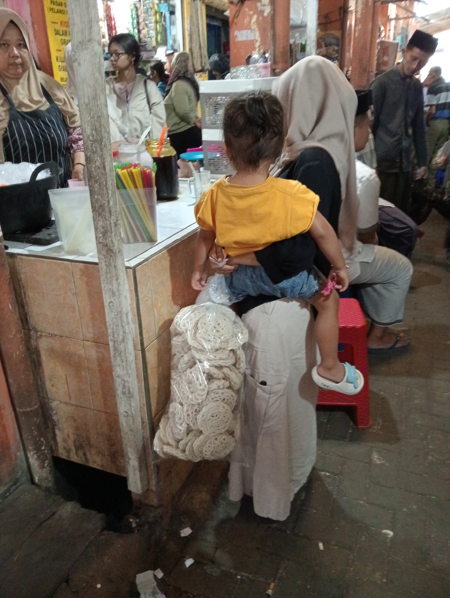 In Grabag, Indonesia, a woman wearing a light beige hijab and a black and white striped apron stands behind a wooden counter. She looks forward, her face partially obscured by the apron. In front of her, a young child with dark hair, wearing a yellow t-shirt and blue shorts, sits on the lap of another person who is mostly out of frame, but wearing a light-colored garment. The child is facing away from the camera, looking down at something in their lap. A bag of white, circular crackers is positioned to the right of the child. Behind the counter, there are containers filled with colorful straws and a dark liquid. The environment appears to be a market or food stall, with wooden structures and tiled surfaces visible.