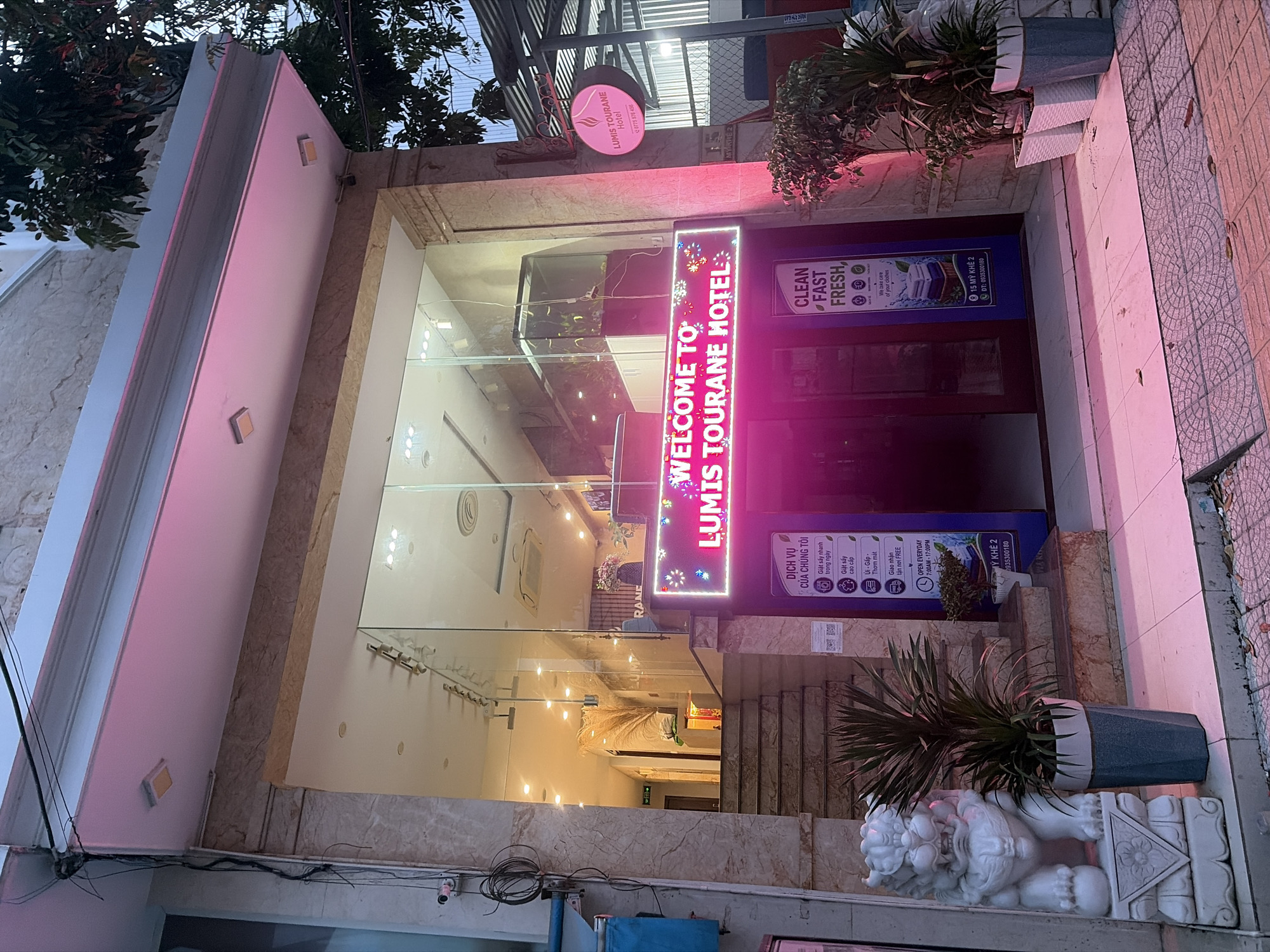 The image shows the exterior of Lumis Tourane Hotel, located at 15 My Khe 2 street in the My An ward of Ngu Hanh Son district, Da Nang, Vietnam. The hotel features a bright pink neon sign above the entrance that reads "WELCOME TO LUMIS TOURANE HOTEL." The entrance is recessed, with glass walls allowing a view into the brightly lit lobby. To the left of the entrance, a white stone lion statue stands guard next to a potted plant. A blue sign board displaying laundry services is visible to the right of the entrance. The building's facade is decorated with marble tiles. The time appears to be dusk or early evening, with artificial lighting illuminating the scene.