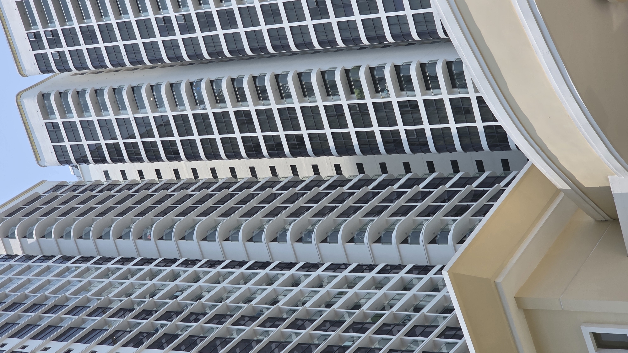 The image shows a section of a high-rise building in Nha Trang, Vietnam. The architecture features a repeating pattern of dark windows set within a white grid. Some windows have curved balcony structures. A portion of a tan-colored adjacent building is visible on the right side of the frame. The sky is visible in the upper left corner.
