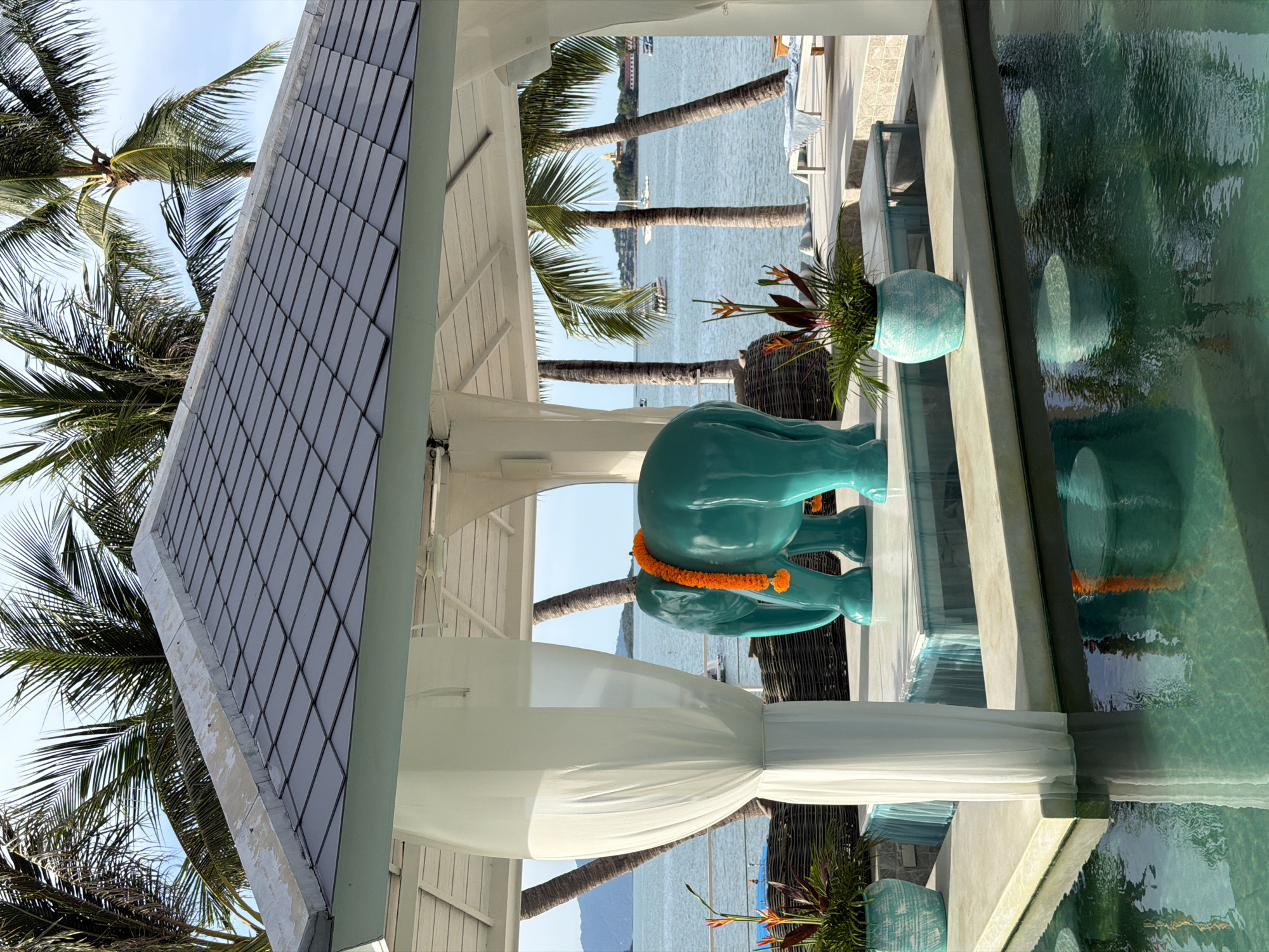 In Baan Bo Phut, Thailand, a turquoise elephant statue adorned with an orange marigold garland stands under a white gazebo. The statue is positioned on a tiered platform with a small blue water feature. White sheer curtains hang from the gazebo, framing the view of the ocean and palm trees. Two potted plants with tropical flowers are placed on either side of the elephant statue. In the background, the ocean is visible with several boats and a distant island. The foreground features a swimming pool with submerged bar stools, reflecting the gazebo and elephant statue. The scene depicts an outdoor seating or relaxation area at a tropical resort.