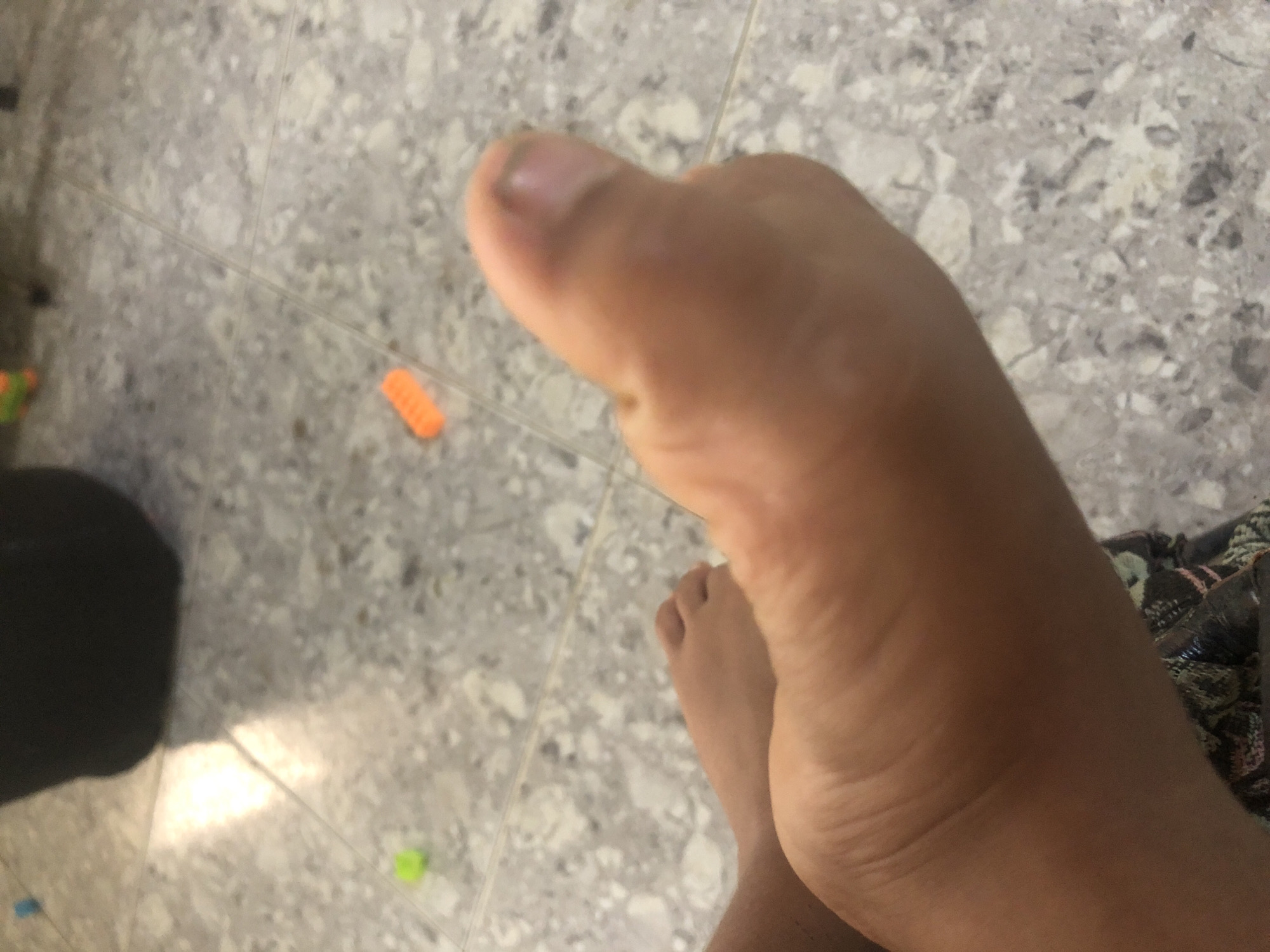 The image shows a close-up view of a person's foot resting on a speckled tile floor. A black object, possibly a bag or piece of furniture, is visible in the background. There are also small, colorful objects scattered on the floor, including a blue and an orange toy. The lighting suggests an indoor setting. There are no discernible landmarks or clues to indicate the location in La Tagua, Venezuela.