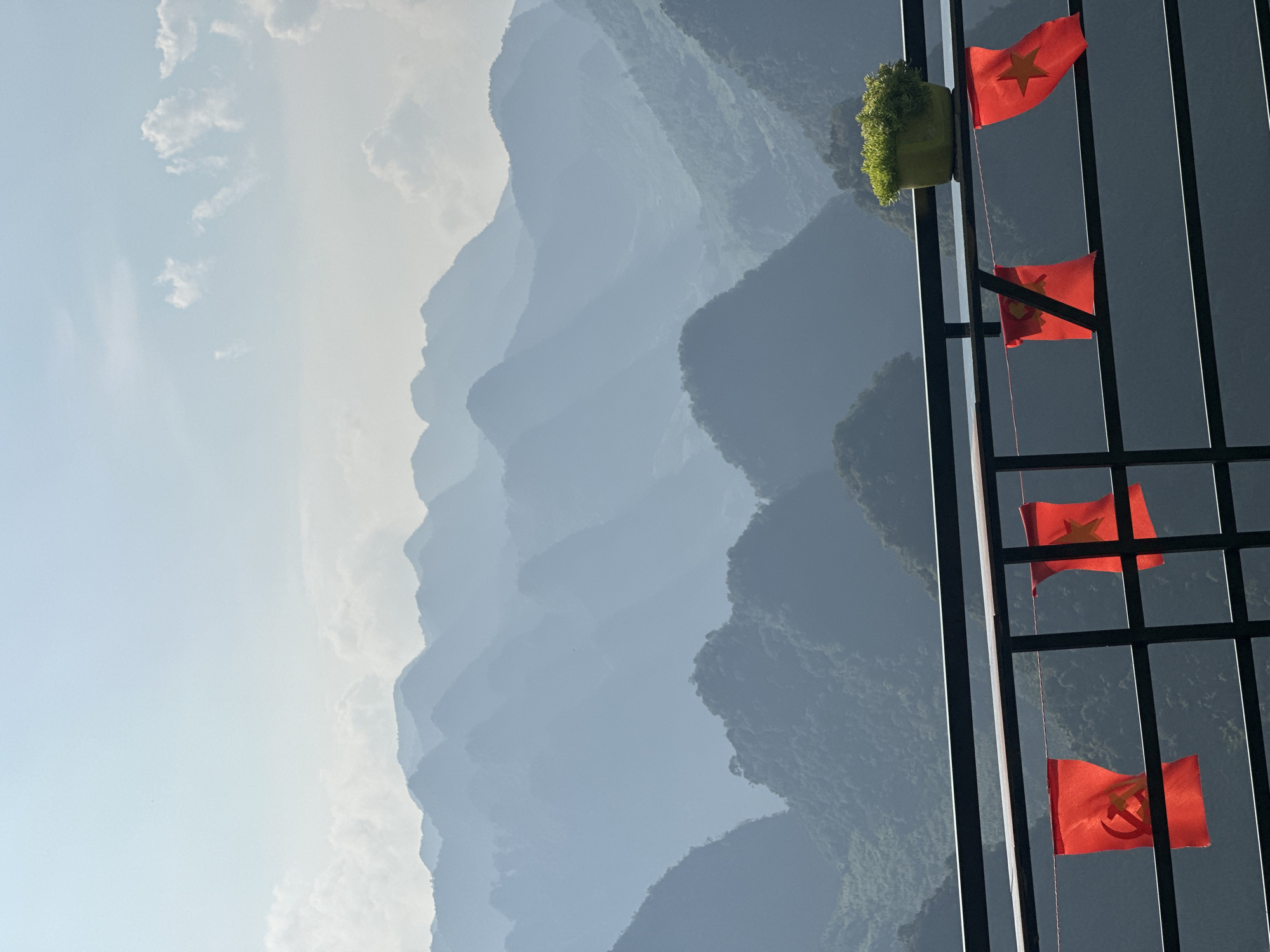 The image shows a view of mountains through a window. Outside the window is a layered mountain range with a hazy, blue-grey color. The sky is visible on the left, with a few white clouds. On the right is a dark, black window frame with a green potted plant on top. There are four Vietnamese flags hanging from the frame. The location is the city of Đường Thượng Commune, Vietnam.