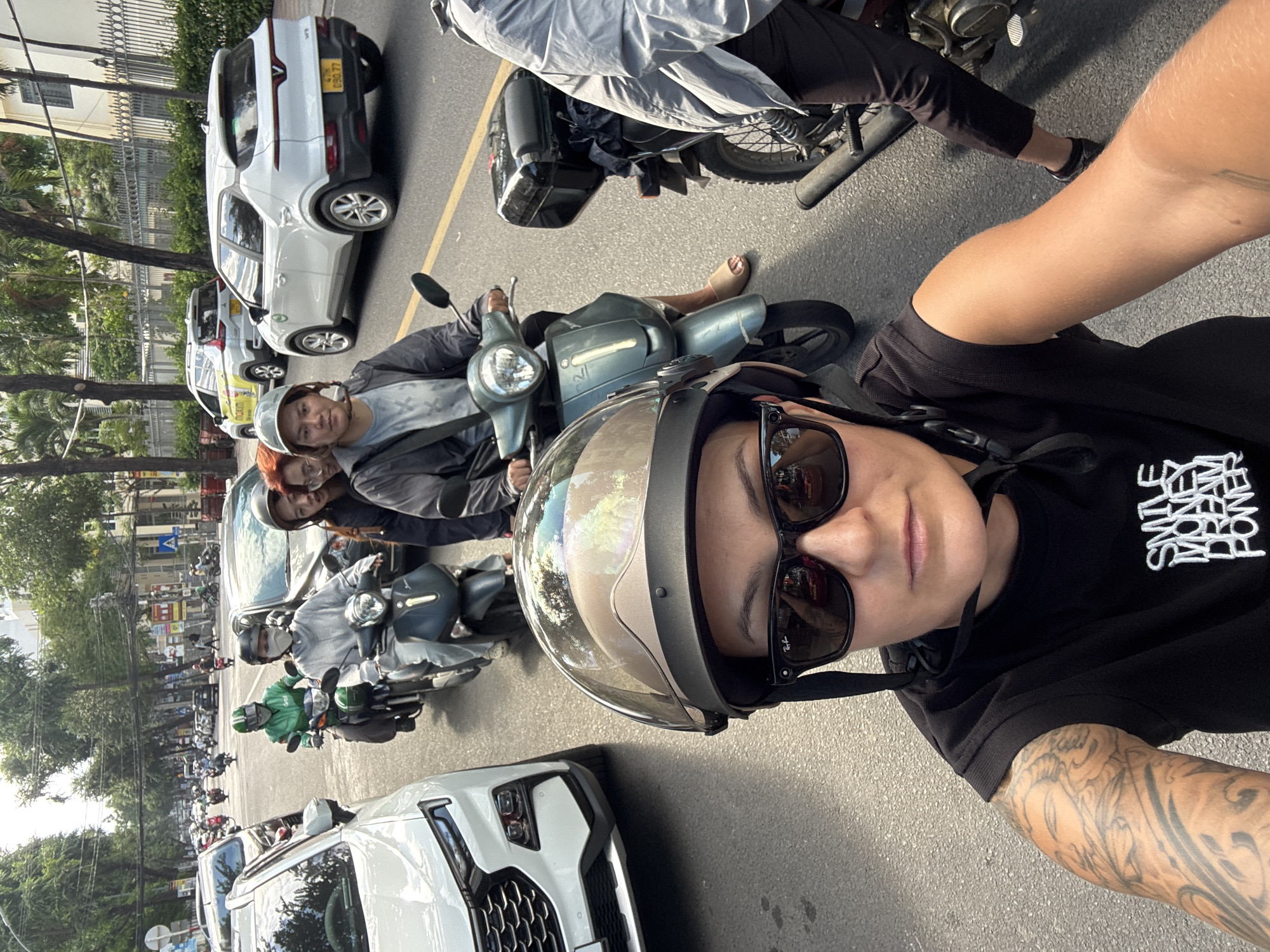 This is a first-person perspective, selfie-style photograph taken from a scooter in moderate traffic on a paved city street in Nha Trang, Vietnam. The street is lined with trees and buildings. In the foreground, a person wearing a black t-shirt with white lettering that reads "SMILE MONEY DREAM POWER", sunglasses, and a helmet with a reflective visor is taking the photo. The person has tattoos on their left arm. To the left of the frame is the front of a white car. Ahead and to the right, other scooters and cars are visible in traffic. One scooter in the middle ground has a rider wearing a green jacket and helmet. Another scooter further back has a rider wearing a grey jacket and a mask. There are several other people and vehicles in the background. The scene appears to be during daylight with sunny conditions, indicated by the shadows and the brightness of the image.
