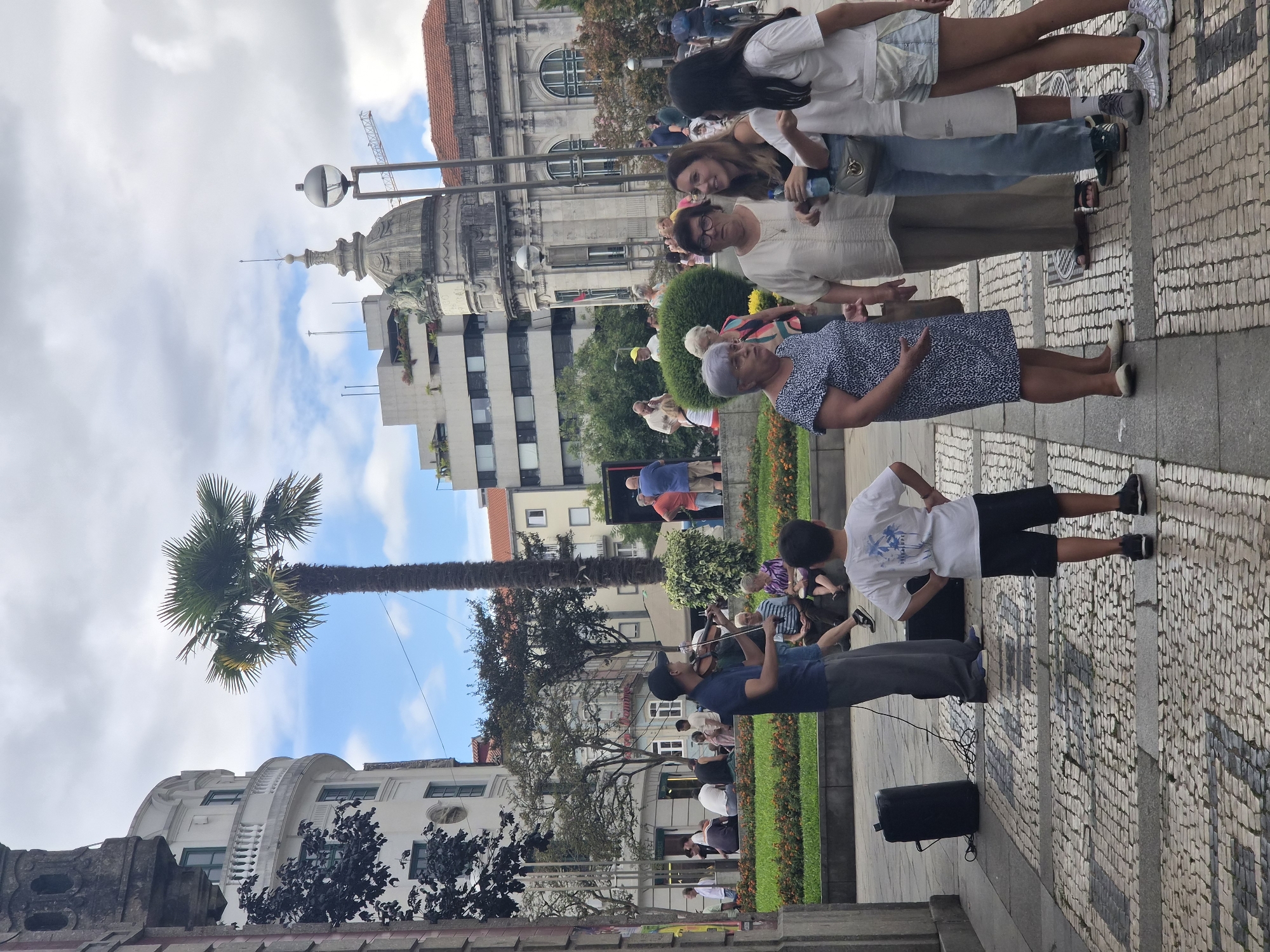 The image captures a street scene in Braga, Portugal, likely in the historic center, given the architectural style of the buildings. In the foreground, a street musician, a man wearing a blue t-shirt and grey pants, plays the violin. He stands to the left of a black speaker and a black amplifier. Behind him, a group of people are seated on a low wall, some watching the performance, others engaged in conversation. A young boy, wearing a white t-shirt with palm trees and black shorts, stands facing away from the camera, observing the scene.
Further into the image, a diverse crowd of people are dispersed on the paved square. To the right, a group of women are talking. One woman, in a blue and white patterned dress, gestures with her hands. Beside her, another woman in a light-colored top holds a water bottle. Next to them, two younger women, one in a white t-shirt and denim shorts, and another in a white top and shorts, are also conversing.
The background is dominated by several buildings. To the left, a white, multi-story building with a rounded facade and balconies is visible. A tall palm tree stands prominently in front of this building. Further back, a more modern, multi-story building with a green roof terrace is present. To the right, a large, ornate building with a dome and classical architectural elements, likely a public building or church, is a focal point. Another building with a red tiled roof is also visible. The sky is partly cloudy with patches of blue. The ground is paved with a mosaic-like pattern of small, light-colored stones. The overall atmosphere suggests a public gathering or a moment of leisure in an urban setting.