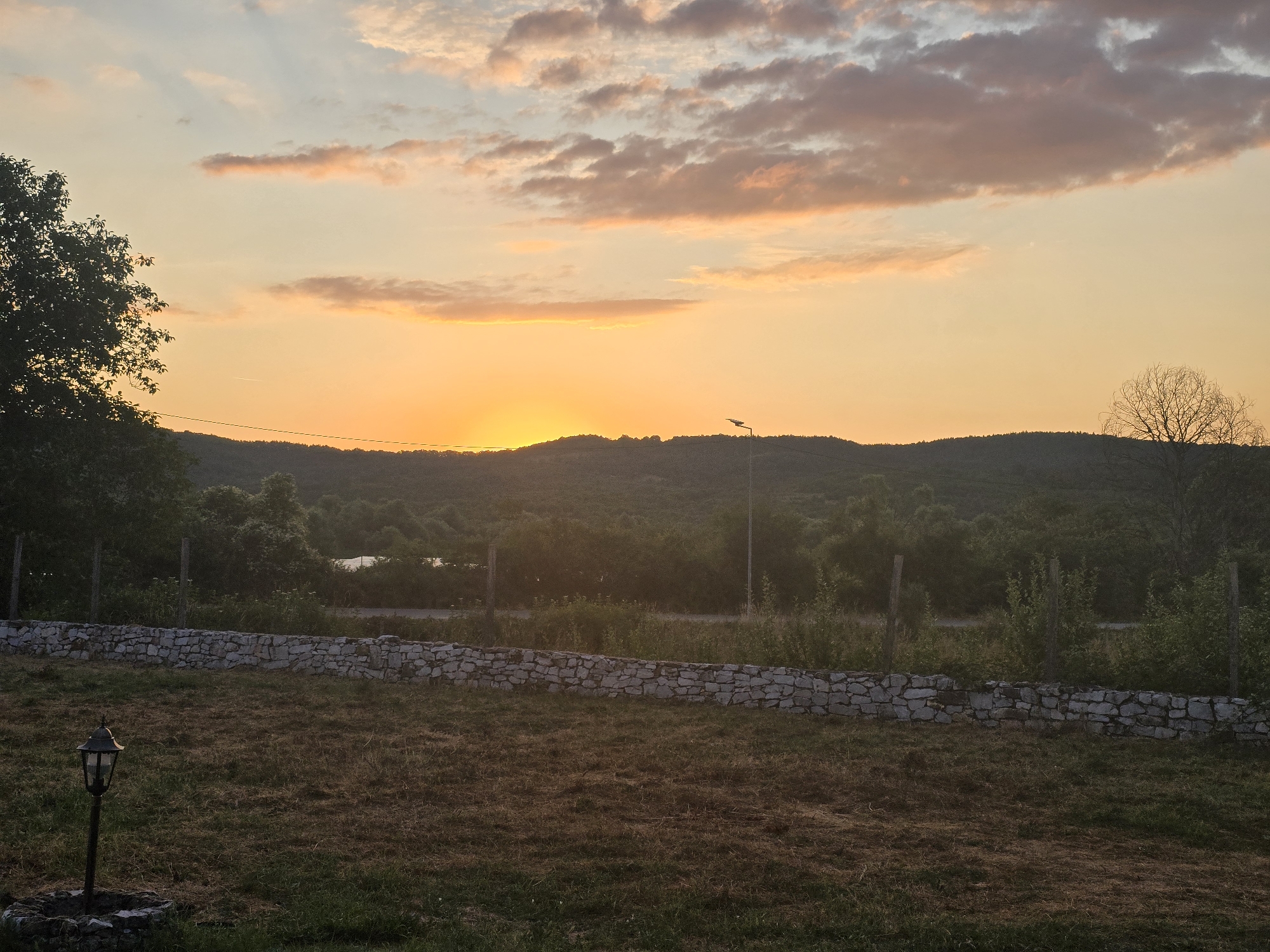This is a wide-angle, low-light, outdoor photograph taken at sunset in the vicinity of Malko Tarnovo, Bulgaria. The scene depicts a rural landscape with rolling hills covered in dense vegetation. In the foreground, a dry, grassy field is divided by a low, stone wall. A vintage street lamp stands in the left portion of the field. The sky is a gradient of warm orange and yellow hues, with scattered, wispy clouds tinged with pink and gray, indicating the setting of the sun. The sun is visible on the horizon, casting a warm glow over the landscape. A single, leafless tree stands silhouetted against the sky on the right side of the image, near the crest of the hills.