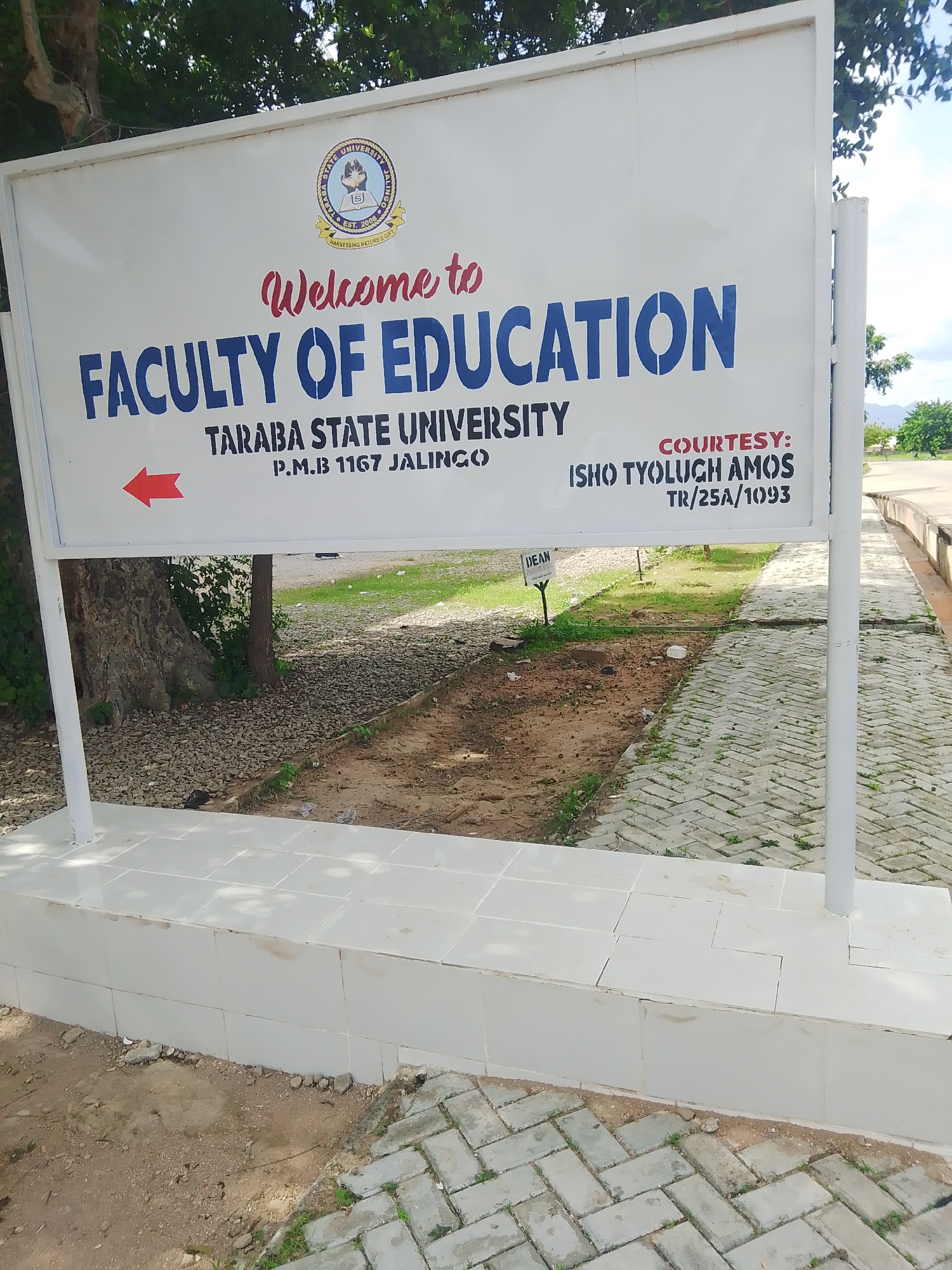 This media file is a photograph of a sign for the Faculty of Education at Taraba State University in Jalingo, Nigeria. The sign is white with blue and red lettering. The top of the sign features the university's logo, which includes the text "Taraba State University," "Est. 2008," and "Harnessing Nature's Gift." Below the logo, the words "Welcome to" are written in red cursive. The main text on the sign reads "FACULTY OF EDUCATION" in large blue block letters. Beneath this, it states "TARABA STATE UNIVERSITY" and "P.M.B 1167 JALINGO." To the left of this text, a red arrow points to the left, indicating direction. On the right side of the sign, under the heading "COURTESY:", the names "ISHO TYOLUGH AMOS" and "TR/25A/1093" are listed in smaller black text. A small sign with the word "DEAN" is visible on a post in the background, to the left.
The sign is mounted on white posts and is situated outdoors. The background shows a tree to the left, a dirt and gravel area, and a paved walkway made of interlocking bricks. In the further background, there is green grass and distant hills or mountains under a bright sky, suggesting it is daytime and likely fair weather. The overall scene depicts an entrance or directional sign for an academic institution.