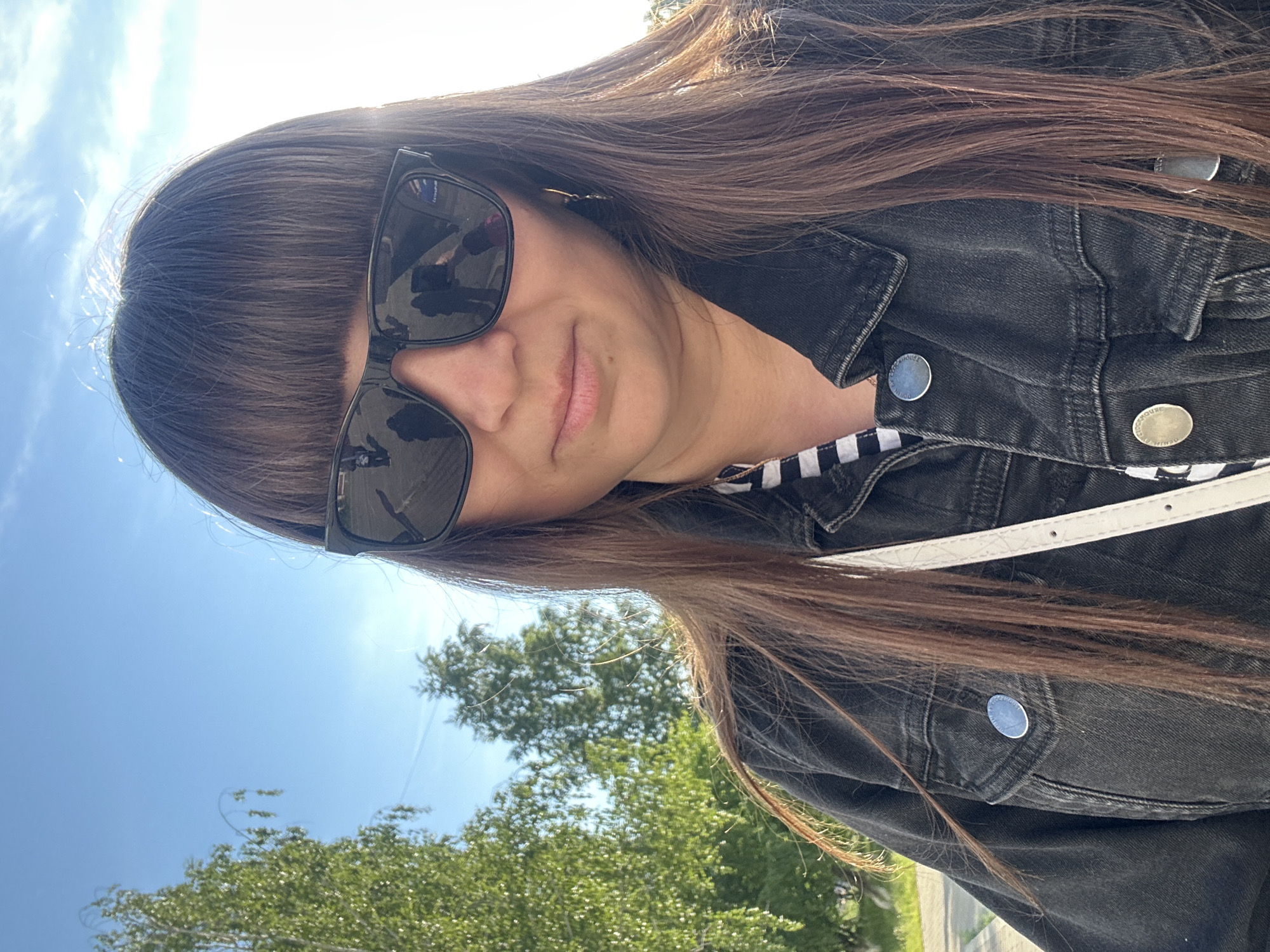 This media file is a close-up, rotated portrait of a person outdoors, captured in Chaikovsky, Russia.
**Subjects:** The primary subject is an individual, likely female, with long, straight dark brown hair styled with bangs. Their face is partially visible behind large, rectangular black sunglasses, through which some reflections can be seen. A faint, closed-mouth smile is visible on their lips.
**Objects:** The person is wearing a black denim jacket with several metallic silver buttons. A striped black and white top is visible beneath the jacket. A white strap, possibly from a shoulder bag or purse, crosses the image over the jacket.
**Scene/Setting:** The background features a clear, bright blue sky with a few wispy clouds. Below the sky, lush green trees are visible, suggesting an outdoor environment such as a park or a general natural area. A light-colored strip in the lower right background might indicate a road or path.
**Activity or event taking place:** The person appears to be posing for a photograph, possibly a selfie, given the close-up and slightly upward angle from which the photo is taken. The overall impression is one of casual outdoor activity.
**Notable details:** The bright lighting indicates it is daytime, likely a sunny and clear day. The image itself is rotated approximately 90 degrees counter-clockwise. Reflections in the sunglasses are subtle but show elements of the surrounding environment.
**Visible text:** On the metallic buttons of the denim jacket, the words "DENIM" and "HOUSE" are discernible, indicating branding.