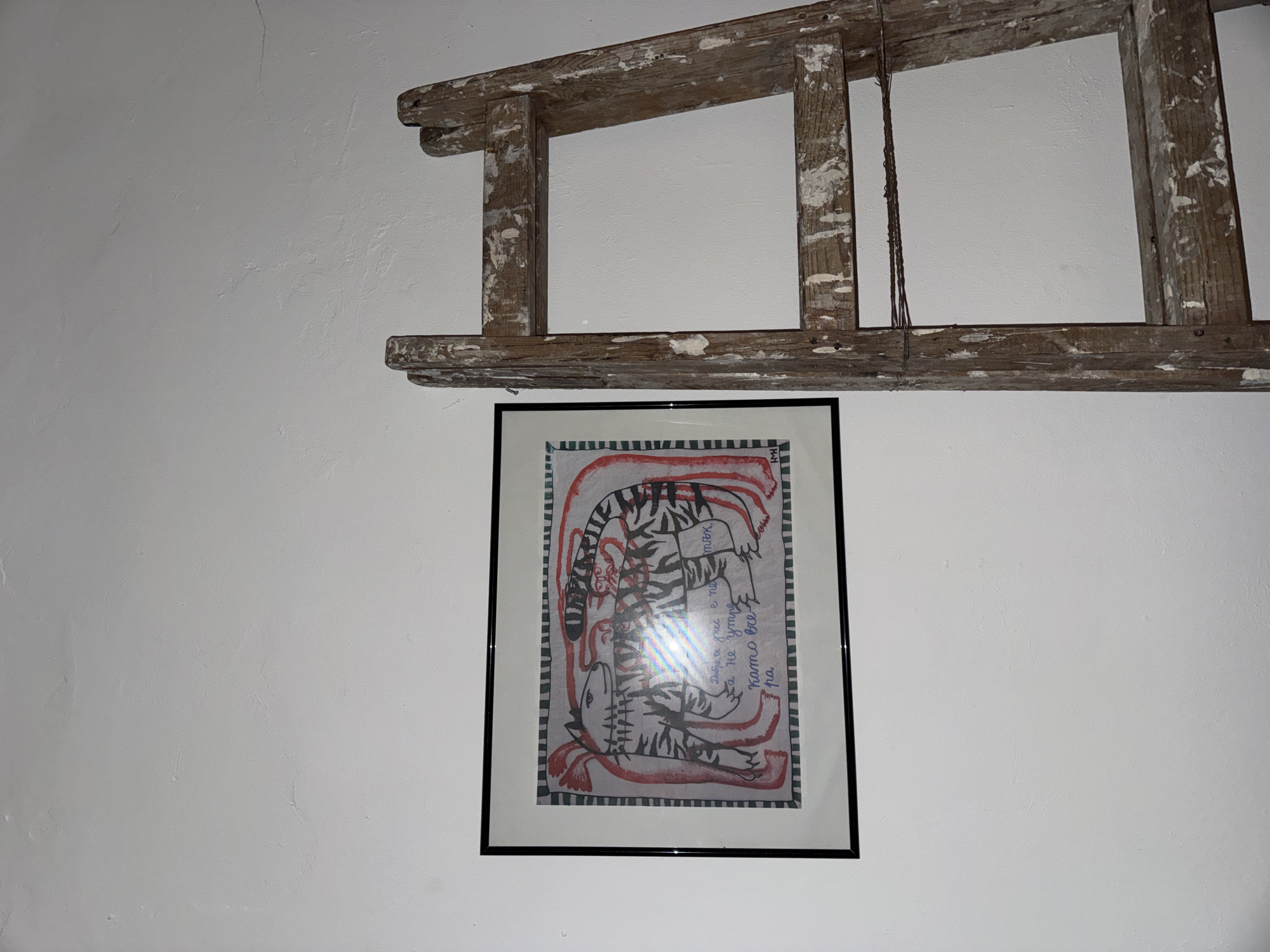 An interior shot displays a white, textured wall adorned with decorative elements. Dominating the upper portion of the frame is an old, weathered wooden ladder, placed horizontally, its rungs and side rails showing significant wear, splintered wood, and remnants of white paint. The ladder appears to be repurposed as a rustic wall decoration.
Below the ladder, centrally positioned on the wall, hangs a framed piece of artwork. The artwork itself is a drawing or painting featuring stylized, striped animal-like figures, possibly tigers, rendered with black stripes and red outlines or accents against a lighter background. The figures are intertwined and abstract. There is a slight glare on the glass of the frame.
Handwritten text, identified by OCR as Bulgarian, is visible within the artwork, integrated into the composition. The text reads: "Добре, че днес е ne / Са не утре / Като все- / pa / тък," which translates roughly to "Good that today is not / But not tomorrow / Like always / pa / so," or a similar fragmented sentiment. A small signature or initial, "HWH", is also visible in the bottom right corner of the drawing. The overall scene suggests a curated, perhaps bohemian or rustic, aesthetic in an indoor setting.