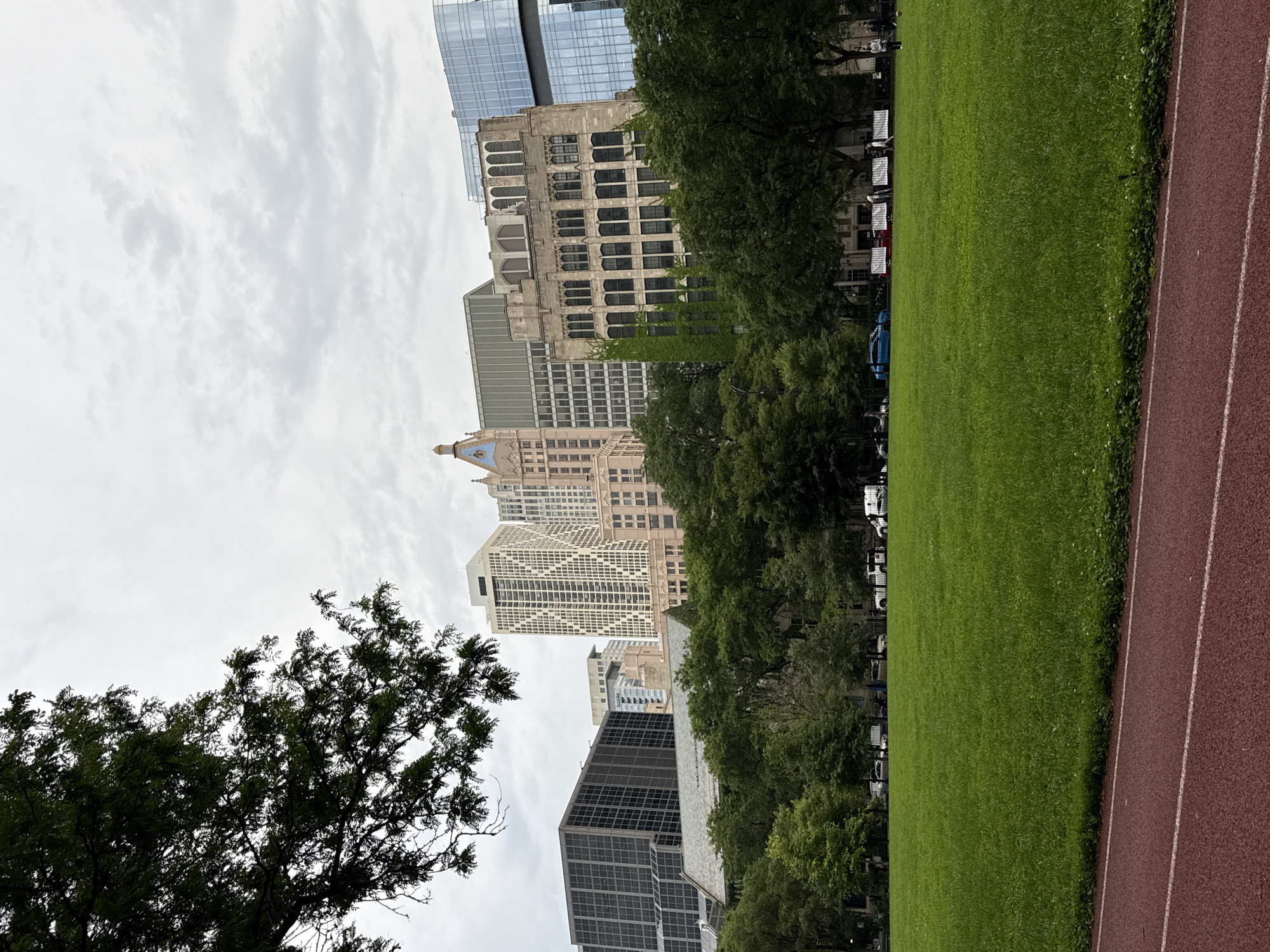 A scenic view of Chicago, Illinois, unfolds with a lush green park in the foreground, featuring a running track with red turf and white lines. Beyond the park, a dense line of mature trees forms a natural barrier, behind which a collection of diverse architectural structures rise. The buildings showcase a blend of styles, from the geometric patterns of a modern skyscraper to the Gothic-inspired arches of an older edifice, with a distinct light pink building featuring a blue spire in the center. The sky above is overcast, with scattered clouds hinting at a potentially temperate day. A few vehicles, including a white van and a bright blue car, are visible on a street bordering the park. Several individuals are scattered throughout the park, some walking, others conversing, and one appears to be kicking a soccer ball, adding a sense of activity to the urban landscape.