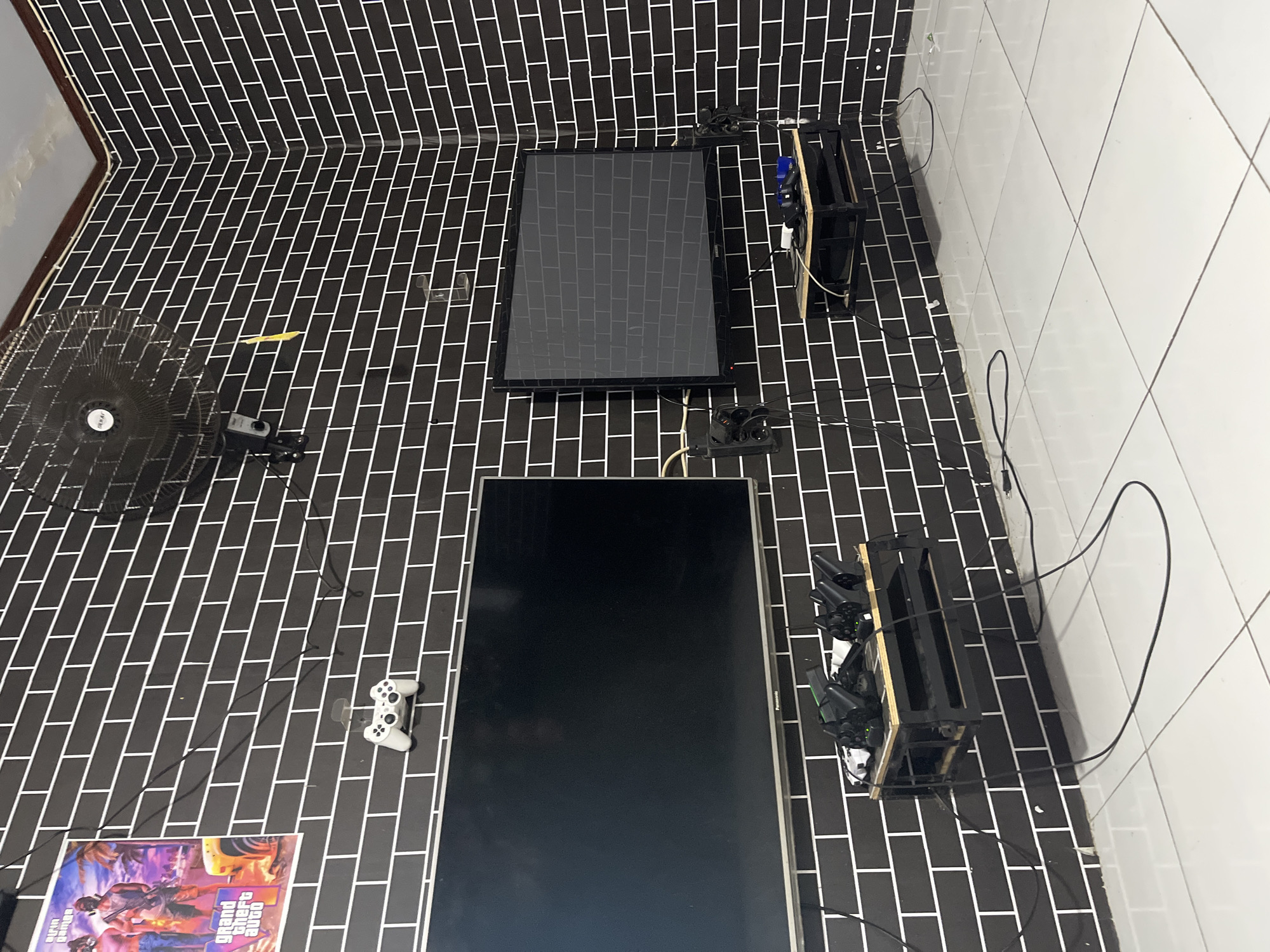 The image depicts a gaming room with a black brick-patterned wall in Laban, Indonesia. Two large televisions are mounted on the wall. The television on the left is black and has a faint reflection. The television on the right is also black and displays a grey brick pattern, indicating it is likely off or in a screensaver mode.
Below the left television, a black metal shelf unit holds several video game controllers, some of which appear to be charging. A white game controller is mounted on the wall above this television.
Below the right television, another black metal shelf unit is present, also holding game controllers and what appears to be a blue game console. Electrical outlets and cables are visible on the wall near the right television.
A poster featuring characters and the "Grand Theft Auto" logo is affixed to the upper left portion of the wall. A black oscillating fan is mounted on the upper right wall. The floor is tiled in white. The overall impression is a dedicated space for playing video games.