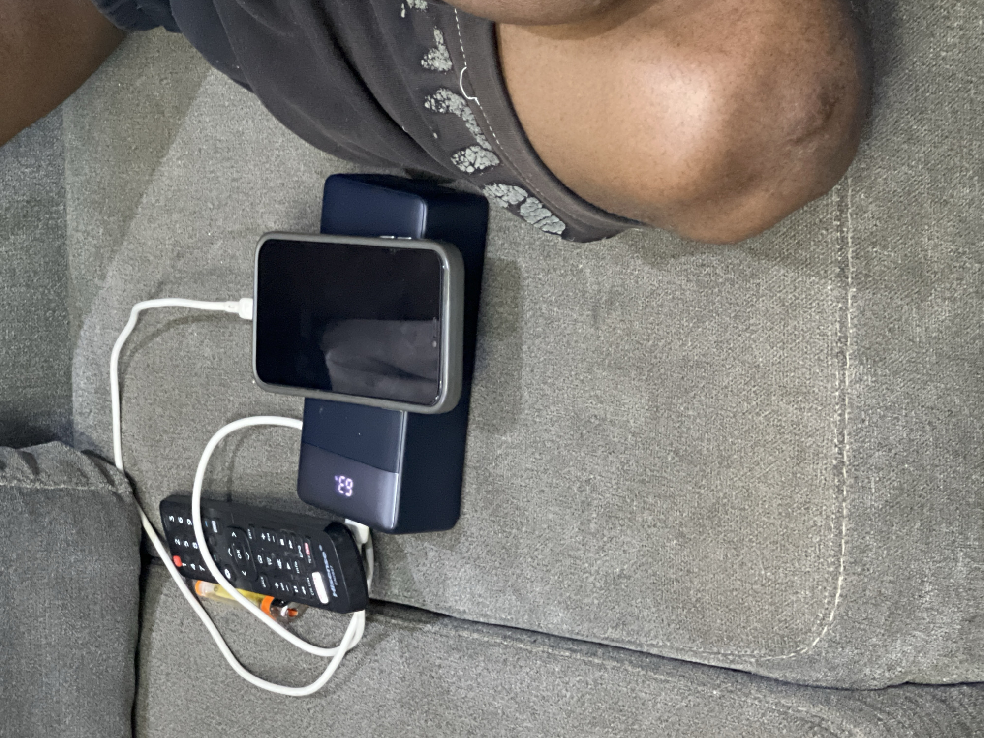 The image captures a close-up, slightly overhead view of a person's leg and knee resting on a textured grey fabric sofa, likely in a living room or lounge area in Abuja, Nigeria. The person is wearing dark shorts or trousers, and their skin tone suggests a person of color.
In the foreground, a dark-colored smartphone with a black screen and a silver/grey frame is placed horizontally on top of a dark blue or grey power bank. A white charging cable connects the smartphone to the power bank, indicating the phone is being charged. The power bank features a digital display showing "63.", which likely represents 63% of its remaining battery charge.
Adjacent to the charging setup, a black television remote control is visible. The brand "Hisense" is clearly printed on it, along with various buttons and numbers. Another white cable, possibly a second charging cable or one meant for the power bank itself, is loosely coiled around the remote control. The overall scene suggests a moment of casual relaxation, with electronic devices being utilized or charged while the person rests on the sofa. The lighting appears to be typical indoor ambient light.