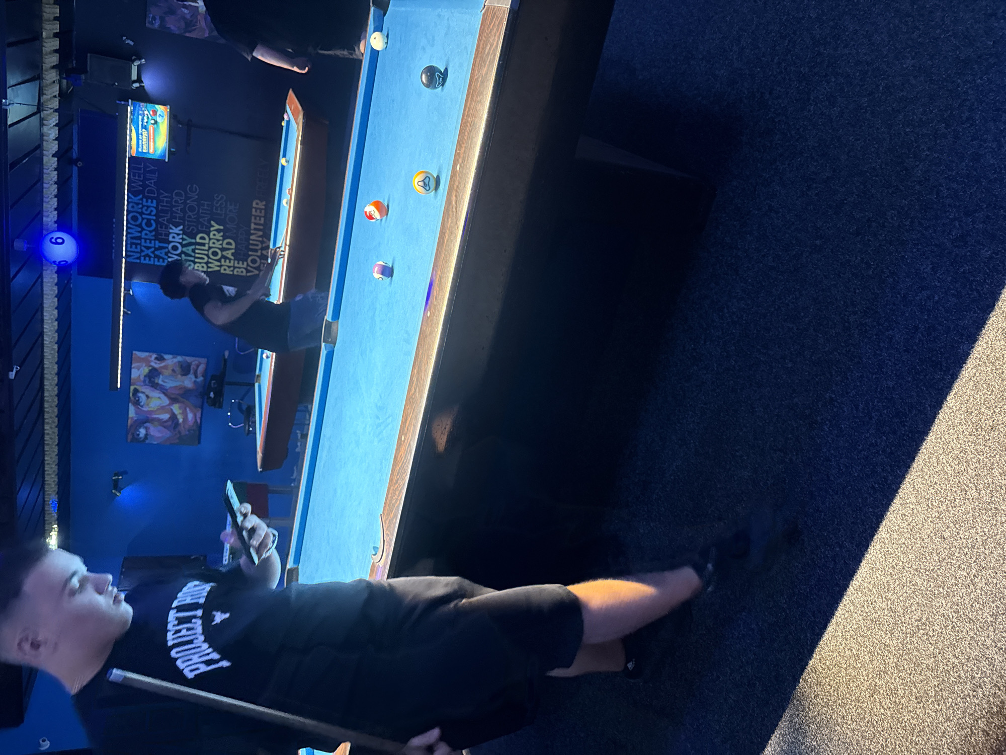 The image depicts a group of people playing pool in an indoor establishment. The scene appears to be a dimly lit room with blue lighting, typical of a bar or entertainment venue. A pool table with blue felt is centrally located, with several billiard balls scattered across its surface. In the foreground, a young man wearing a black t-shirt with the text "PROJECT H" is holding a mobile phone and a pool cue, seemingly distracted by his device. To the right, another person in a dark shirt is playing pool, while a third individual, partially visible, is also present. The background features a wall with motivational text and artwork, including a painting and a screen displaying a game. A spherical light fixture with the number "6" illuminated is suspended from the ceiling. The floor is covered in a dark blue carpet. Without further information, it is impossible to determine the specific location of this establishment within Yambol, Bulgaria.