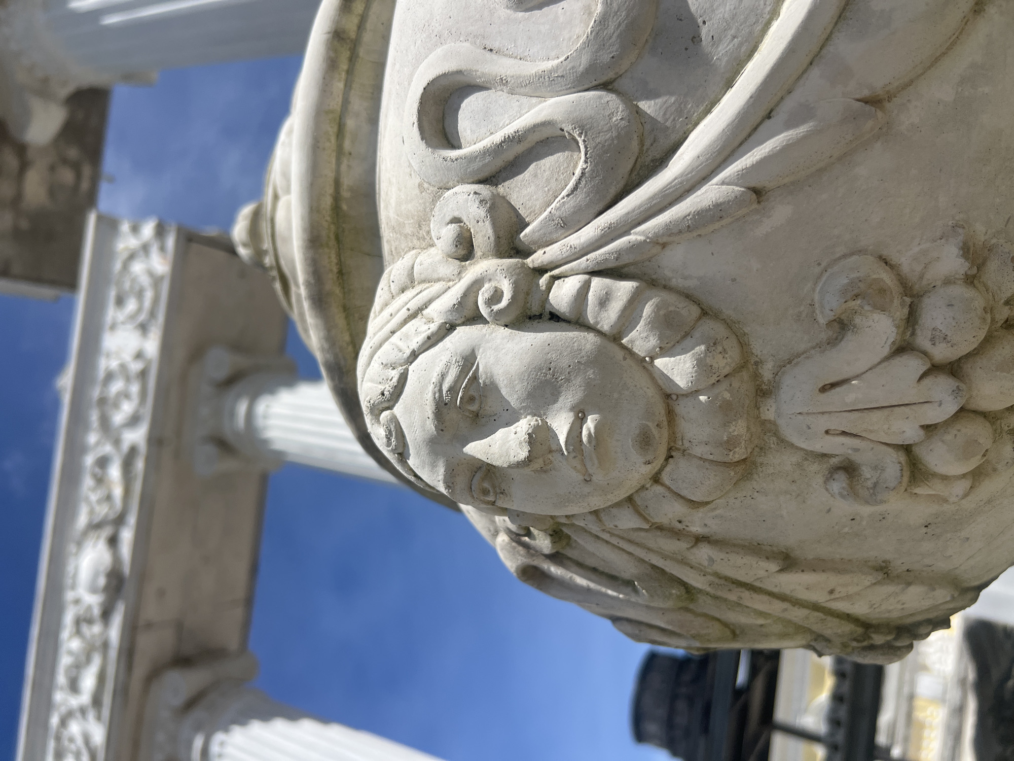 A close-up, low-angle shot focuses on a sculpted face on the side of a white, stone urn. The urn is ornate, featuring carved hair with curls and swirling decorative elements. The face itself has a serious expression with distinct features such as eyes, a nose, and lips. Behind the urn, a bright blue sky is visible, with the tops of white classical columns and a decorative architectural frieze blurred in the background. The overall scene suggests an outdoor setting, possibly a garden or a historical site in Pa Khlok, Thailand, given the architectural style and clear sky.