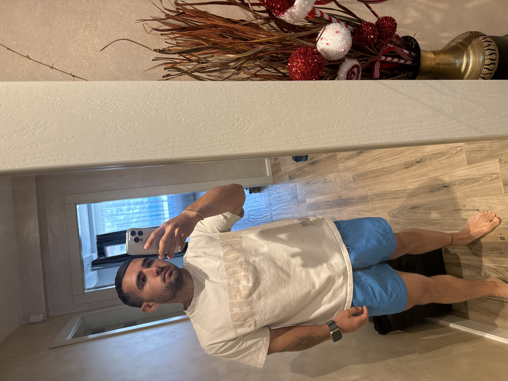 A young man with short dark hair and a beard takes a mirror selfie in what appears to be a residential hallway. He is wearing a white t-shirt with a beige logo and bright blue shorts. A smartwatch is visible on his left wrist. To the right of the frame, a vase with decorative reeds and red and white ornaments, possibly for a holiday, is visible. The floor is made of light-colored wood-look tiles. The background shows a doorway leading to a room with a window and curtains. There is no visible indication of the specific location in Gabrovo, Bulgaria.
