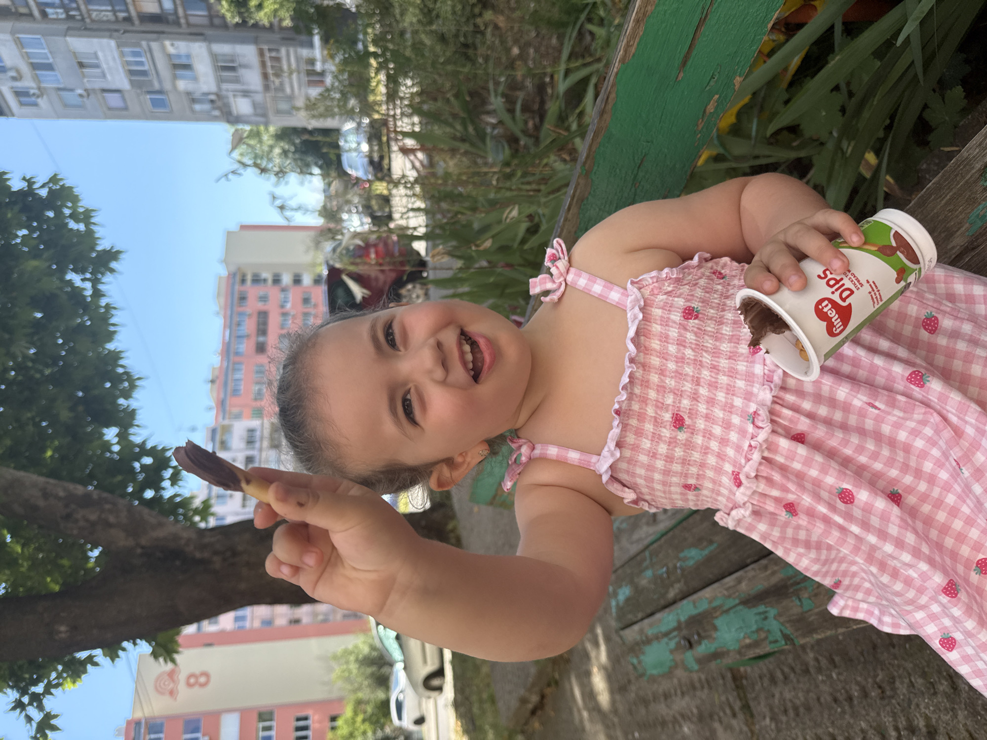 A young girl with brown hair, wearing a pink checkered dress with a strawberry pattern and white ruffled trim, sits on a weathered green bench in an outdoor setting. She is holding a stick dipped in chocolate in her right hand and a cup labeled "fineti Dips" in her left hand. The cup contains chocolate dip and small, crunchy pieces. The background features trees, apartment buildings, and parked cars, suggesting an urban environment. The scene is lit by natural daylight under a clear blue sky. The location is identified as Burgas, Bulgaria.