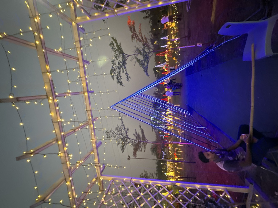 An outdoor scene in Kalyan-Dombivli, India, depicts an illuminated evening event or festival space. The foreground features a large white metal pergola or archway overhead, heavily draped with warm-yellow string lights that also cascade downwards. To the left, a young adult male is seated on a dark stool, holding a light-colored bamboo stick, wearing a dark, patterned short-sleeved shirt and dark trousers. A light blue plastic chair is partially visible to his right.
Central to the image is a triangular tunnel-like structure made from multiple parallel blue-glowing LED strips, creating an illuminated passageway. A dark blue carpet or mat covers the ground within this tunnel. Beyond this structure, an expansive field or park area is densely populated with various illuminated decorations, including red and white conical lights, numerous yellow ground lights, and other abstract glowing installations. Several individuals are observed walking along a path further into this brightly lit background. Two distinct trees with sparse foliage are silhouetted against a pale, desaturated blue-grey sky, indicative of twilight or early evening conditions. The ground beneath the structures and in the immediate background appears to be reddish-brown earth or grass.