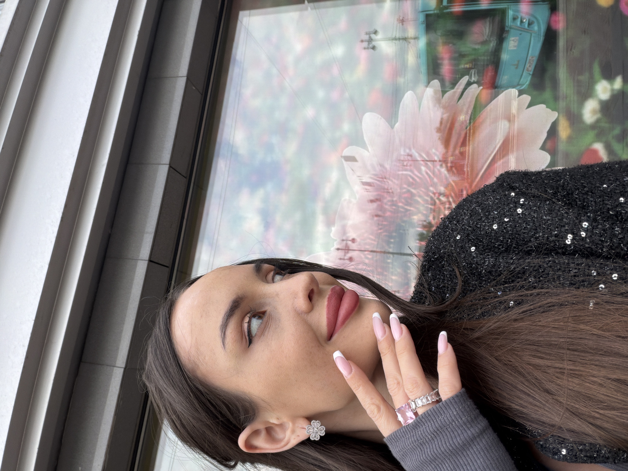 The image is an eye-level portrait of a woman with long brown hair, wearing a black sequined top and a grey long-sleeved shirt. She has fair skin, light brown eyes, and is wearing makeup, including lipstick and well-manicured nails with a white French tip. She is also wearing a flower-shaped earring and a ring with a large pink stone and a row of square-cut diamonds.
The woman is positioned to the right side of the frame, looking upwards and to the left. Her hand is raised, with her fingers slightly spread apart, displaying the ring.
The background consists of a building exterior with vertical grey panels on the left and a large window reflecting a colorful flower arrangement and the silhouette of a vintage blue bus or vehicle. The reflection also seems to include power lines and streetlights, typical of an urban environment.
Based on the appearance of the bus reflected in the window, the woman's style, and the overall design of the scene, there's a possible association with the designer brand Alexander Terekhov, which is based in Saint Petersburg. It is implied that the location could be in the city of Saint Petersburg, Russia, due to the prevalence of the fashion house there.