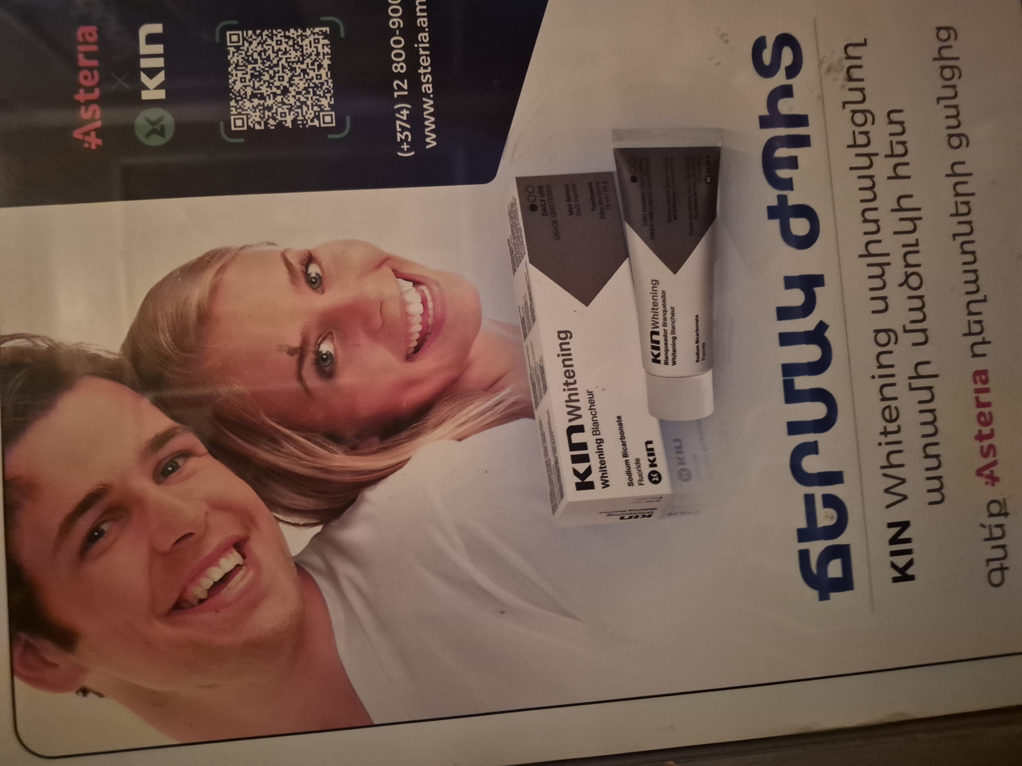 The image is an advertisement featuring a young, smiling Caucasian couple. The woman has blonde hair and a small mole on her forehead. They are both looking towards the viewer, with the man on the left and the woman on the right. In the foreground, there are two tubes of "KIN Whitening" toothpaste, one partially visible on top of another. The packaging of the toothpaste is white with dark grey accents and text in English and French.
Below the toothpaste, there is text in Armenian. The large text reads "ԲԵՐԱՆԻ ԺՊԻՏ," which translates to "Smile of the Mouth" or "Smile of the Lips." The smaller text below that says "KIN Whitening սպիտակեցնող ատամի մածուկի հետ," meaning "KIN Whitening with whitening toothpaste." The final line of text at the bottom reads "զևէք Asteria դեղատների ցանցից," which translates to "from the Asteria pharmacies network."
The upper right corner of the advertisement features the "Asteria" logo and the "KIN" logo. A QR code is also visible. Below the QR code, contact information is provided: "(+374) 12 800-900" and "www.asteria.am." The country code "+374" indicates that the advertisement is for a business located in Armenia, and given the context of "Asteria pharmacies," it is likely an advertisement displayed in or associated with a pharmacy in Yerevan, Armenia. The overall environment is that of a print advertisement, with a light background for the couple and a dark background for the brand information.