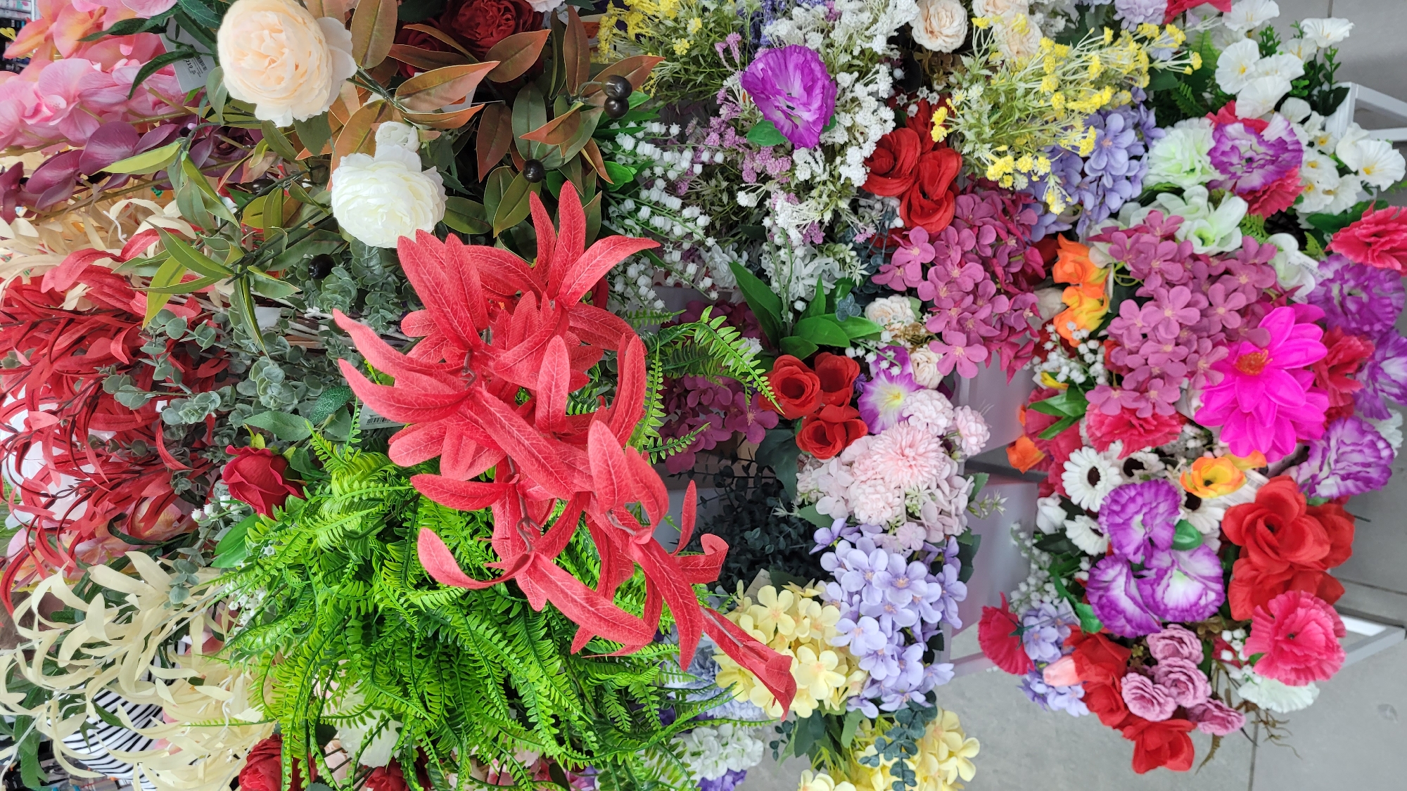 A high-angle, medium shot depicts a dense arrangement of artificial flowers of various colors, types, and sizes. Dominating the center-left are vivid red, spiky artificial flowers interspersed with green ferns and other foliage. Adjacent to these are white and peach-colored roses, interspersed with red and brown leaves. The right side of the image is filled with a mixture of colorful flowers, including purple, pink, white, red, yellow, and orange blossoms. Some of these appear to be hydrangeas and daisies. The background and specific location are indeterminate. No people are visible.