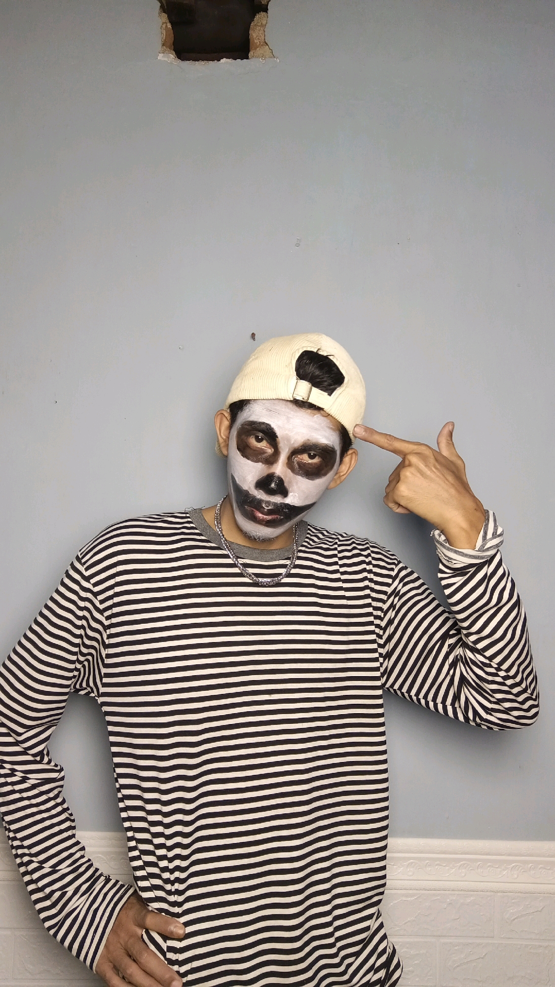 A person wearing a striped shirt and a baseball cap is posing for a photo against a light blue wall. The person has skull makeup on their face, with white paint covering their face and black paint around their eyes and nose. Their right hand is raised to their head, with their index finger pointing towards their temple, mimicking a gun shape. The person is looking directly at the camera. The wall behind them has a large, rough hole near the top. The floor area is not clearly visible, but there appears to be some white decorative molding at the bottom. The overall impression is of a posed, perhaps playful, photograph.
