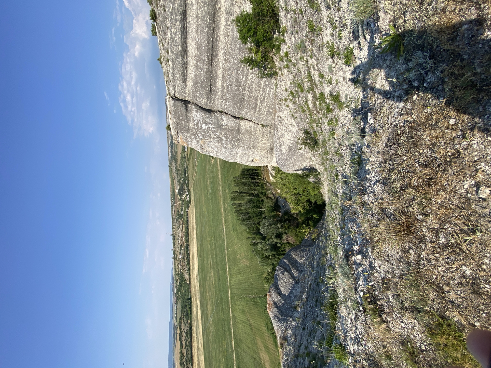 This media file captures a dramatic landscape view from a cliff edge. The scene is dominated by the rugged, grey rock face of the cliff, which shows significant weathering and a deep crack running through it. Small patches of hardy green vegetation cling to the rock, suggesting a dry, sunny environment.
Below the cliff, a vibrant green agricultural field stretches out, marked by faint lines suggesting cultivation. A darker band of trees borders the field, creating a contrast with the lighter green. In the distance, the land rolls gently, with hints of more vegetation and structures. The sky is a clear, bright blue, with a few wispy white clouds scattered near the horizon, indicating fair weather.
The perspective is from a high vantage point, looking down and out over the landscape. A shadow of a person is visible on the rocky ground in the foreground, with their hand or arm also visible at the edge of the frame, suggesting the image was taken by someone standing on the cliff. The lighting and long shadows indicate it is likely late afternoon or early morning. The location cues, including the agricultural field and rugged terrain, combined with the provided location context, point to a rural setting in Ukraine. There is no visible text in the image.