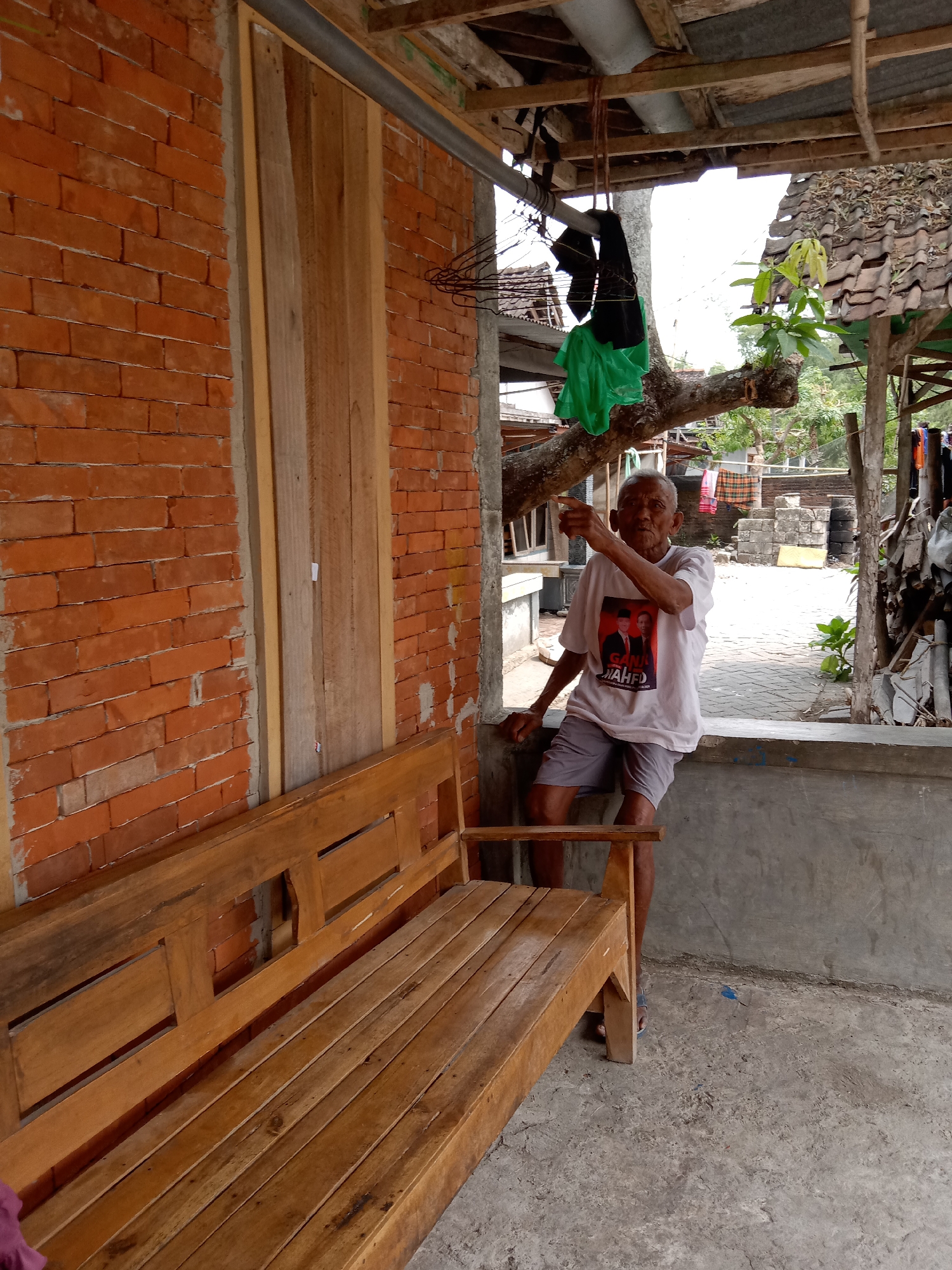 In the city of Jumput, Indonesia, an elderly man stands near a wooden bench, gesturing with his right hand. He wears a white t-shirt with a printed image of two men and text, along with grey shorts. The man is positioned on the right side of the frame, leaning against a concrete ledge. To his left, a wooden bench extends into the foreground. Behind the bench is a red brick wall, and to the right, a concrete wall and paved ground are visible. Overhead, a rustic roof structure with exposed wooden beams and a tiled roof is present, with some clothing items hanging from a branch. The environment appears to be an outdoor area, possibly a residential yard or a communal space.