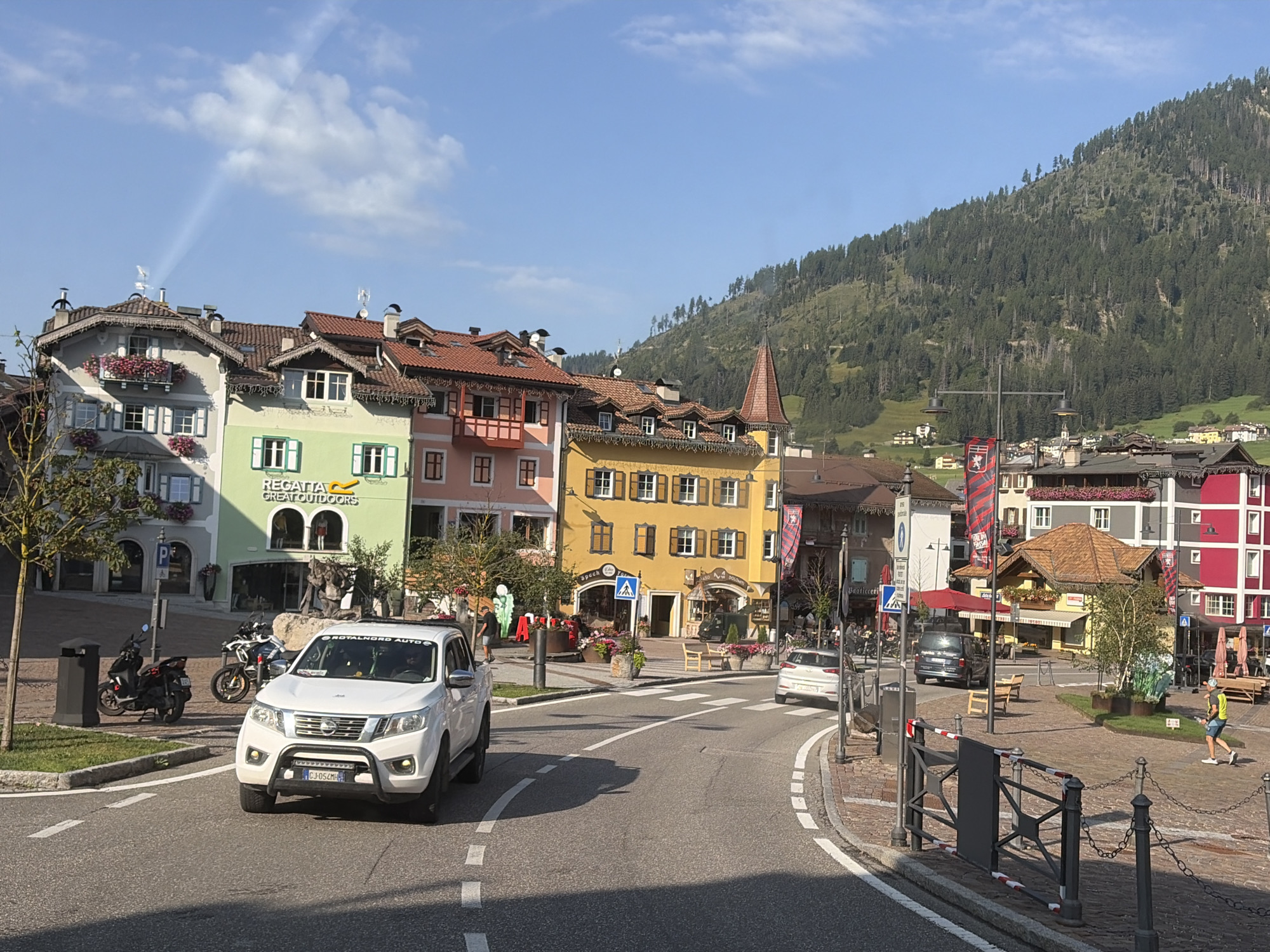 The image shows a street scene in Moena, Italy. In the foreground, a white pickup truck drives on a road. To the left of the road, there are two parked motorcycles and a black trash bin. Behind the trash bin, a tree with a thin trunk is visible. In the middle ground, several colorful buildings with traditional alpine architecture line the street. Some buildings have balconies adorned with flowers. Signs for "REGATTA GREAT OUTDOORS" and "SPECK DOLOMITI" are visible on the facades. Pedestrians are seen walking on the sidewalks and in a plaza. A white car is parked in the plaza. In the background, a steep, tree-covered mountain rises against a clear blue sky with some scattered clouds. The sun casts shadows, indicating it is daytime. The overall scene depicts a charming European town square.