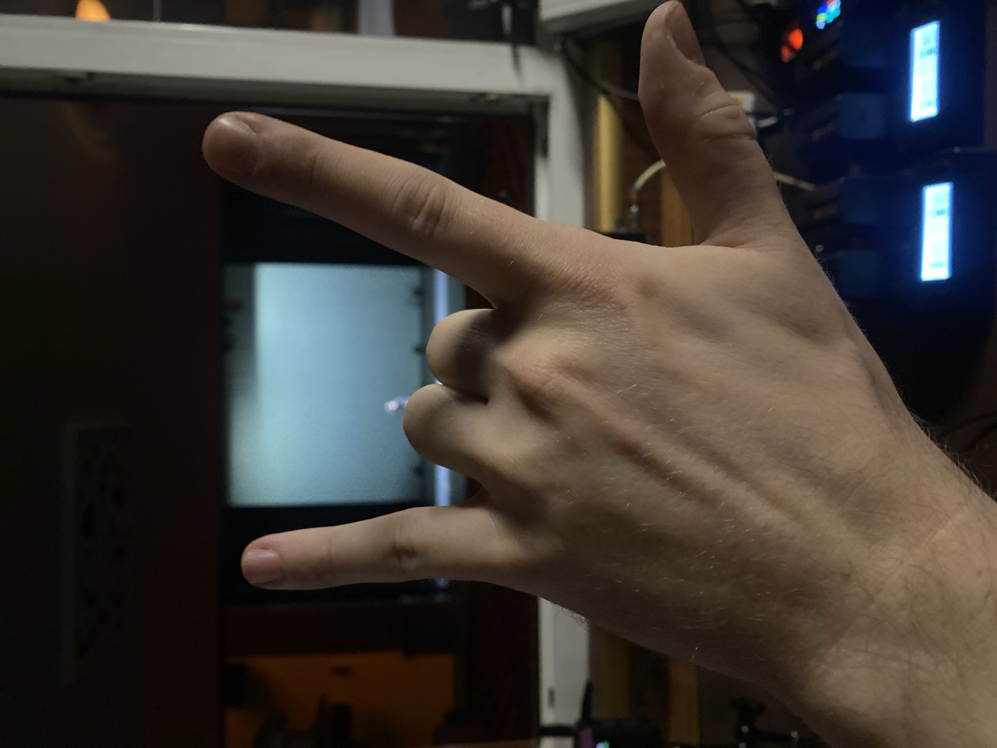 A close-up image displays a Caucasian person's hand in the "rock on" hand gesture. The index finger and pinky finger are extended upwards, while the middle and ring fingers are folded down into the palm, held down by the thumb. The hand is light-skinned and covered with fine hair. The background is out of focus and dark, but shows elements of what appears to be a 3D printer, with screens displaying illuminated text. The location is not determinable from the image, so I cannot provide a location in Rusanovo, Russia.