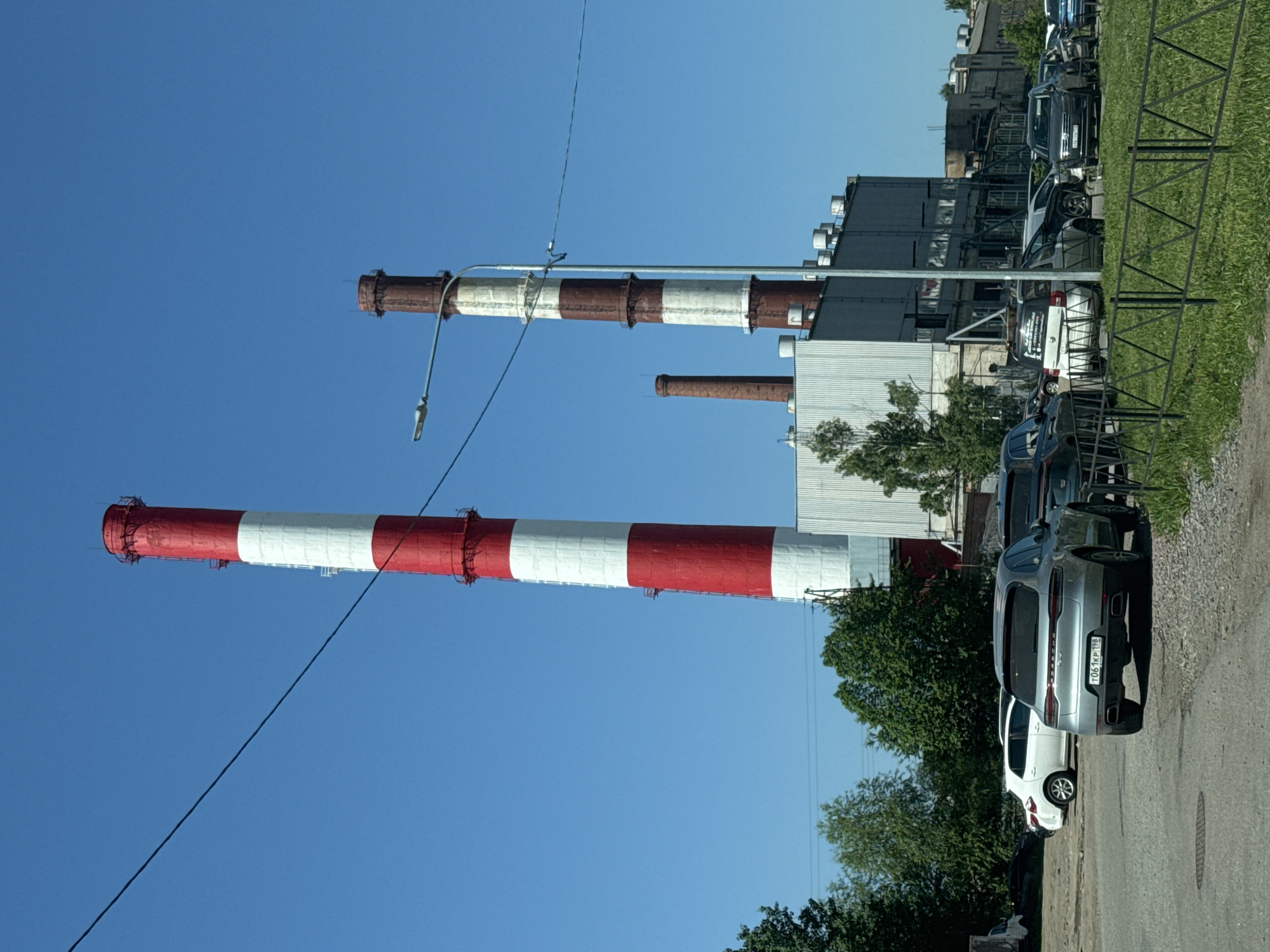 The image depicts an industrial or commercial area in Sinyavino, Russia, under a clear, bright blue sky. Two prominent industrial chimneys, painted with alternating red and white stripes, dominate the left side of the frame, extending vertically. The taller chimney, positioned more to the left, has a street light fixture attached to it, with power lines stretching across the sky. A smaller, plain brick chimney is visible further back, to the right of the taller striped one.
In the background, a series of light-colored industrial buildings with flat roofs and multiple vents are visible. A row of vehicles is parked along a road on the right side of the image. The most clearly identifiable vehicle is a silver Porsche Cayenne SUV, parked facing away from the viewer. Its Russian license plate, "Т061КР 98", is visible on the rear, with "98" denoting the Saint Petersburg region. A white hatchback is parked in front of the Porsche, and other dark-colored cars and SUVs are also present in the parking area.
A black metal fence separates the paved or gravel road from a strip of green grass on the far right. Trees and green foliage are scattered throughout the scene, providing a natural element against the industrial structures. The bright, cloudless sky indicates a sunny daytime setting, likely midday or early afternoon. No people are visible, and the scene is static, showing an overview of the industrial landscape and parked vehicles.