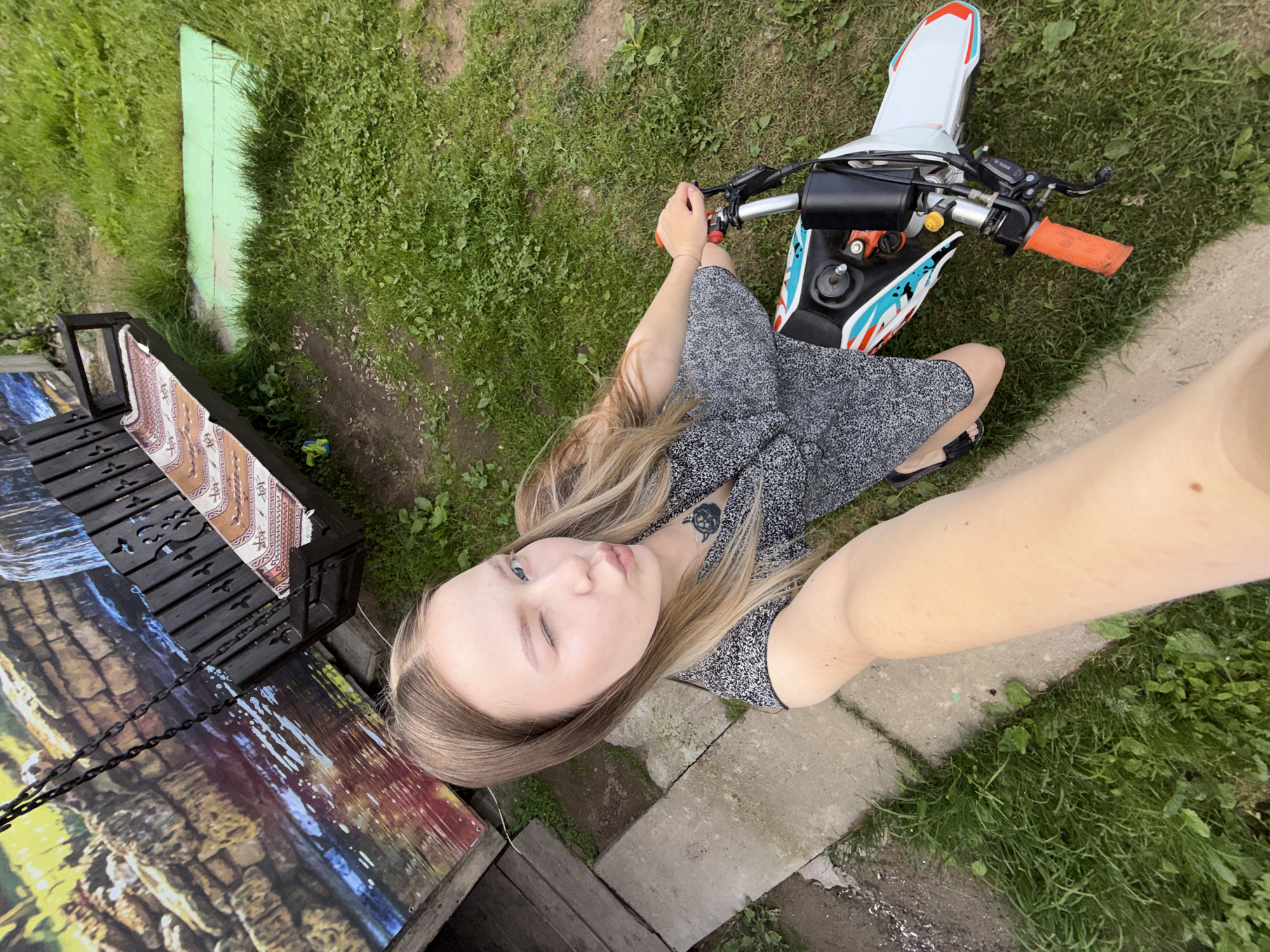 A young woman with long blonde hair is taking a selfie outdoors, sitting on a dirt path next to a dirt bike. She is wearing a short-sleeved black and white speckled dress and black sandals. She is looking at the camera with one eye closed and her lips pursed, giving a playful expression. Her left arm is extended towards the camera, capturing the selfie.
The dirt bike is white with orange handlebars and colorful graphics in red, white, and blue. It is parked on the grass. To the left of the woman, there is a dark wooden bench with decorative cutouts, covered with a patterned fabric and a large, colorful picture of a rocky landscape.
The setting appears to be a rural or backyard area, with lush green grass and some dirt paths. There is a light green wooden plank lying on the grass in the background. The overall atmosphere suggests a casual, sunny day, likely in the afternoon, based on the lighting. The image is taken from a high-angle perspective, looking down. There is a tattoo visible on the woman's neck, resembling a black silhouette of an animal. No visible text is present in the image. The location is identified as Zhigalki, Russia.