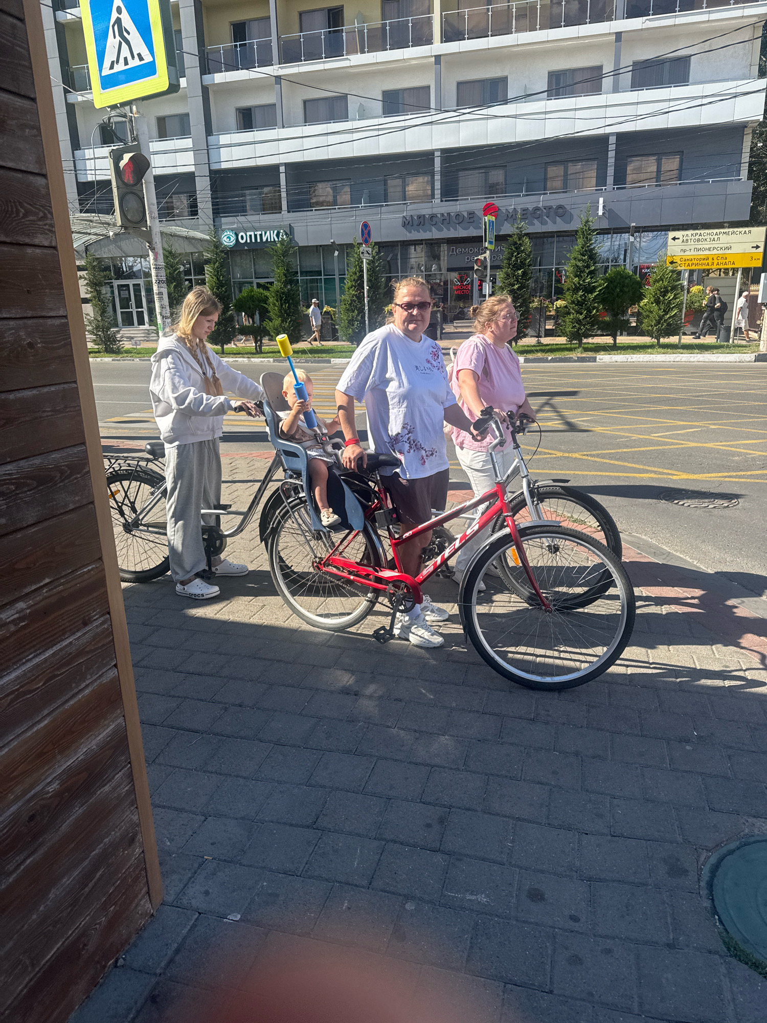 In Anapa, Russia, a blonde woman with braided hair is pushing a bicycle equipped with a child seat on a sidewalk paved with grey interlocking bricks. A toddler, wearing a light-colored shirt and holding a yellow toy, sits in the child seat. Another woman, wearing glasses and a pink t-shirt, is walking next to her, holding the handlebars of a red bicycle. The background features a multi-story building with balconies and shops on the ground floor, including a sign for "оптика" (optics) and "МЯСНОЕ МЕСТО" (meat place). Traffic lights and street signs are visible on the side of the street. Trees line the area in front of the building. The overall lighting suggests it is a sunny day.