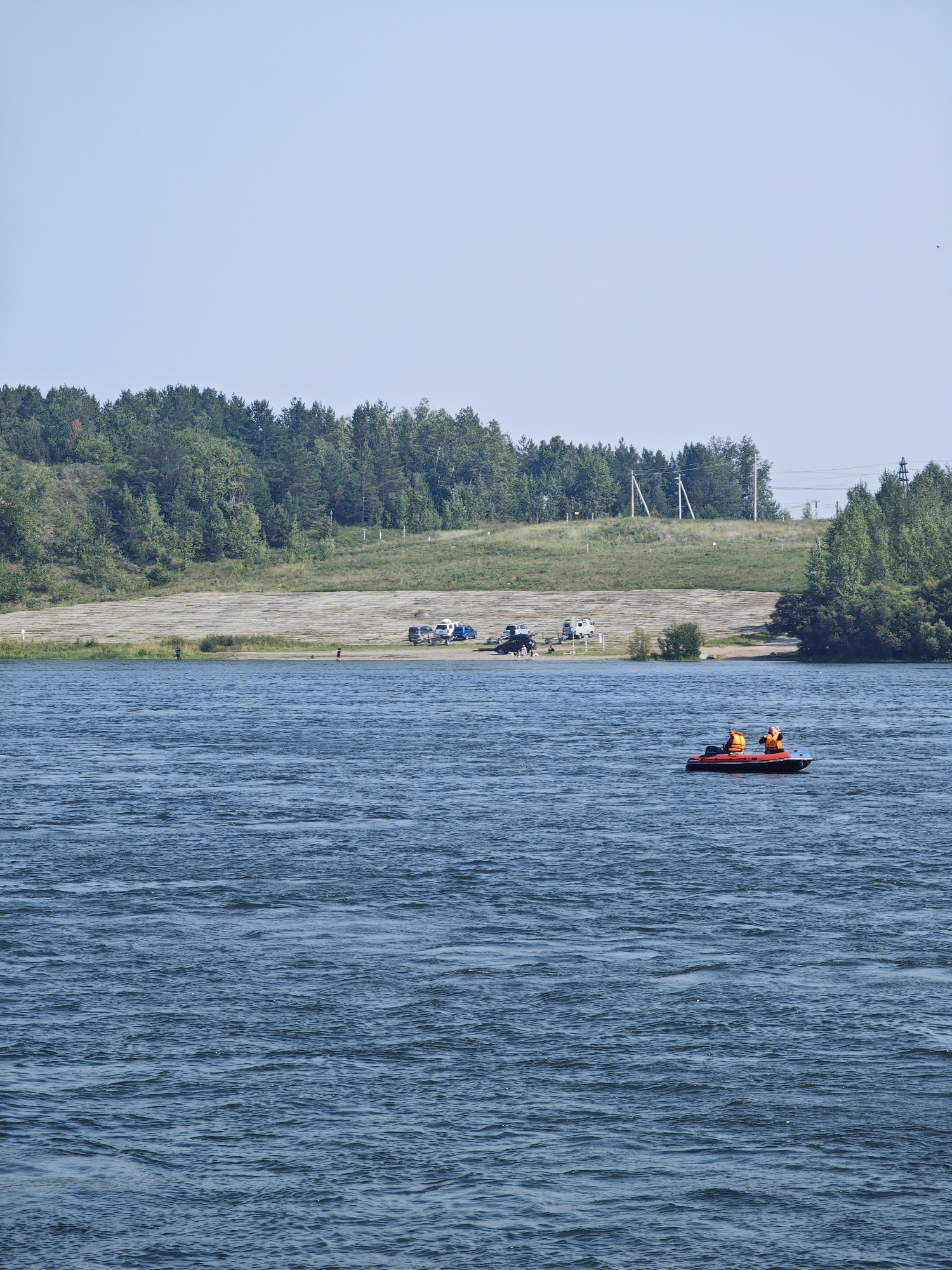 On a clear, sunny day, a wide expanse of blue water fills the foreground of the image. In the middle ground, a small, red and black boat navigates the water, carrying two individuals wearing orange life jackets. To the left of the boat, the shore is visible, featuring a sandy beach area with several parked vehicles, including a white camper van and a blue van. Further inland, a gentle slope is covered in green grass and trees. In the background, a dense line of dark green trees stretches across the horizon, with a clear blue sky above. Power lines and poles are visible on the right side of the frame, suggesting the area is not entirely remote. The overall scene depicts a recreational area by a body of water, likely a lake or reservoir, where people are enjoying outdoor activities. This specific location is likely the Krasnoyarsk Sea, a large reservoir on the Yenisei River, near the city of Krasnoyarsk, Russia.